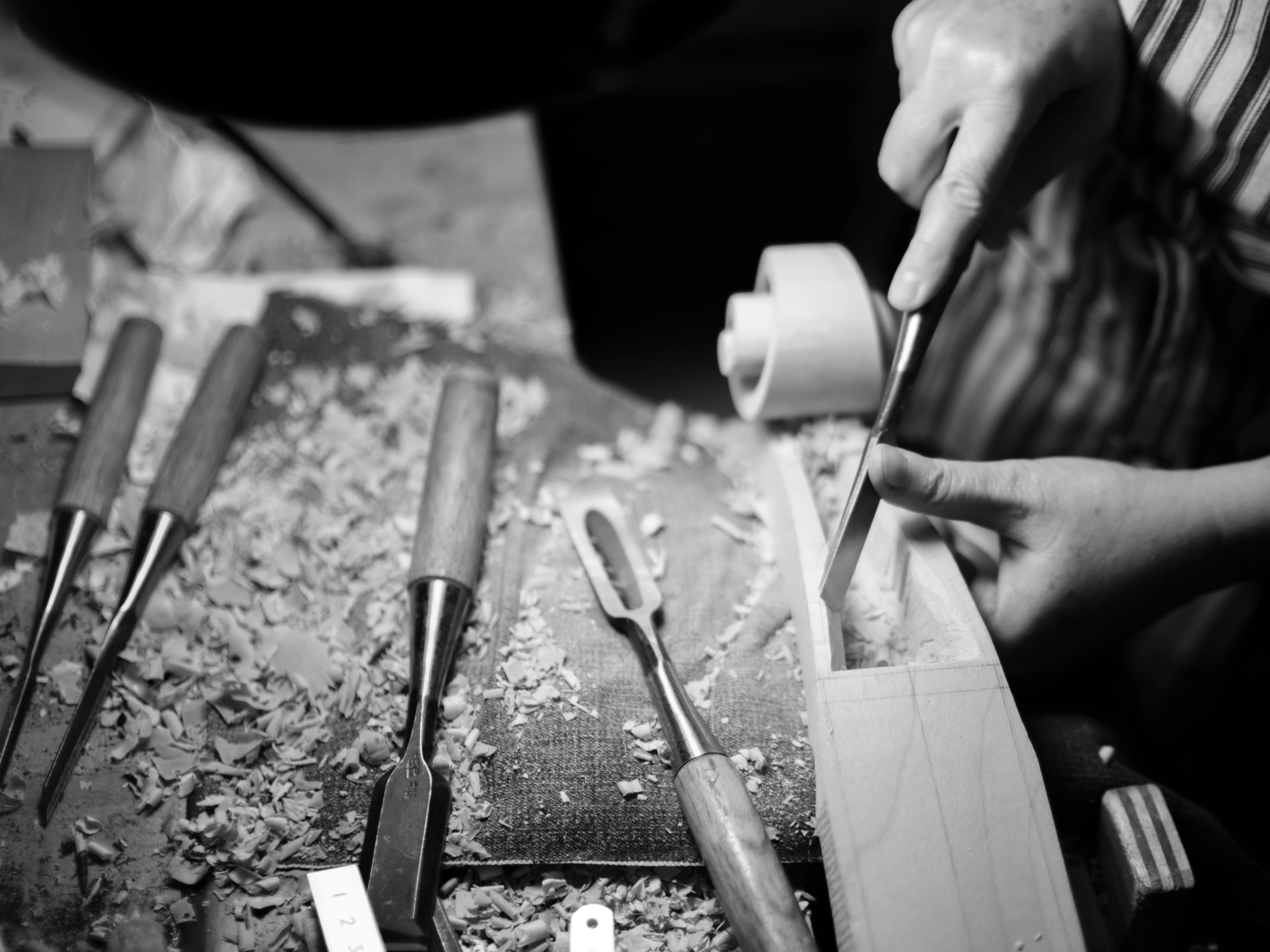 Close up, black and white photo of a hand using a chisel to carve the scroll of a violin on a workbench.