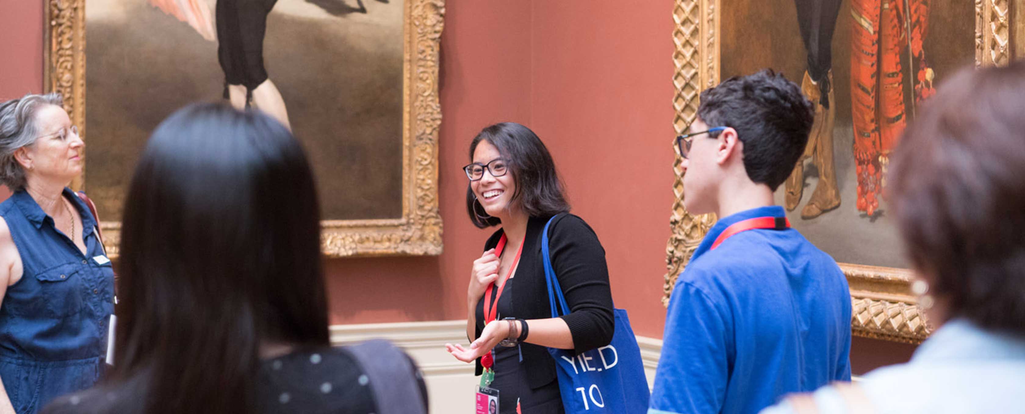 A young woman with long, brown hair, wearing glasses, is smiling as she leads a group of people in a gallery tour.