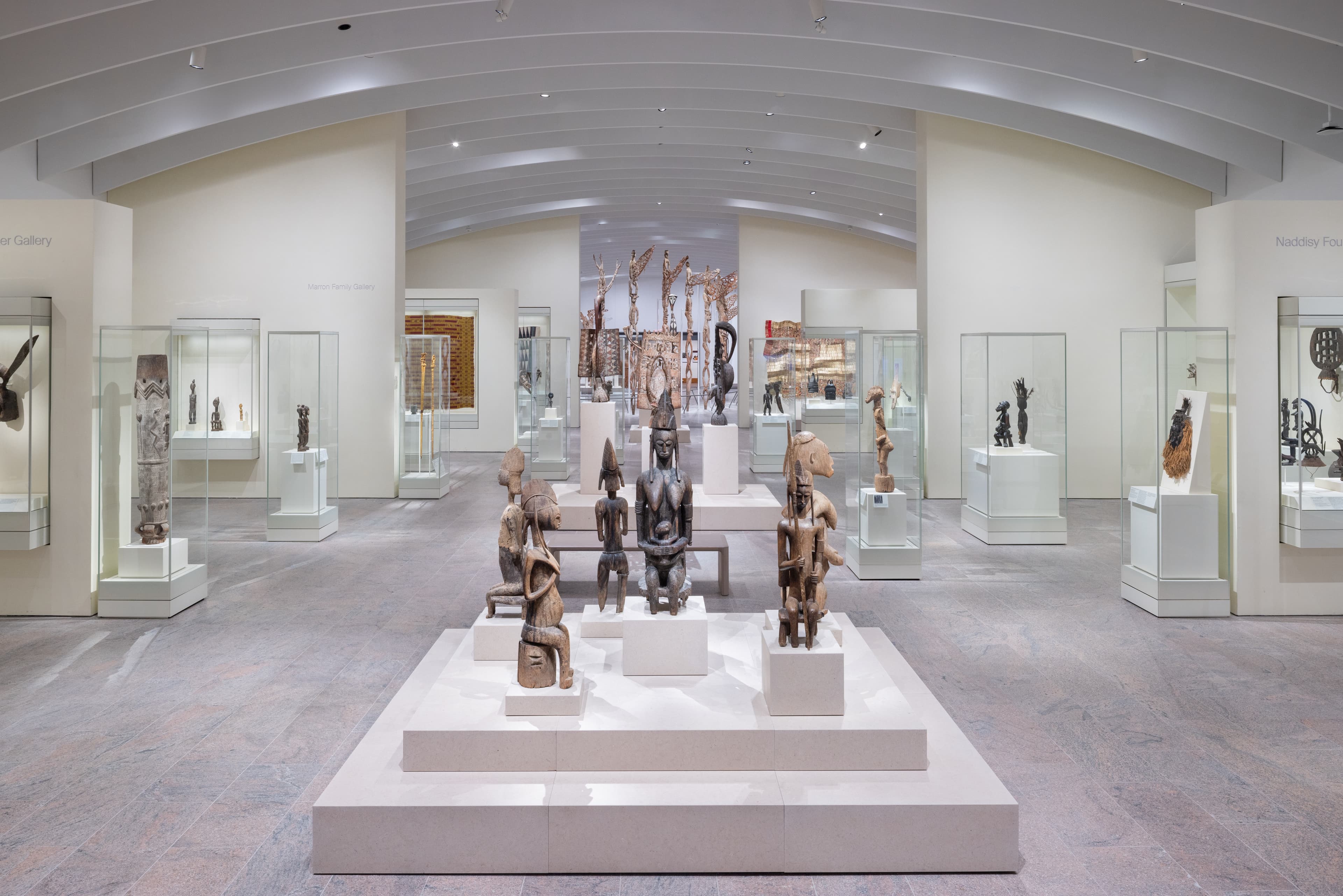 A photograph of the new Michael C. Rockefeller Wing galleries which features a group of sculptures made out of wood in the center foreground and cases with objects inside on the left and right.
