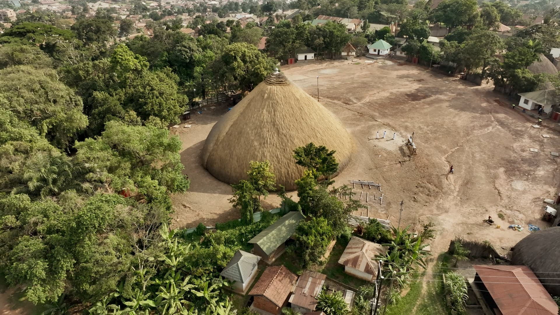 An overhead view of the Kasubi Tombs in Kampala, Uganda. The woven, wattle and daub structure is surrounded by a forest on one side, and homes on the other with an empty lot directly in front.