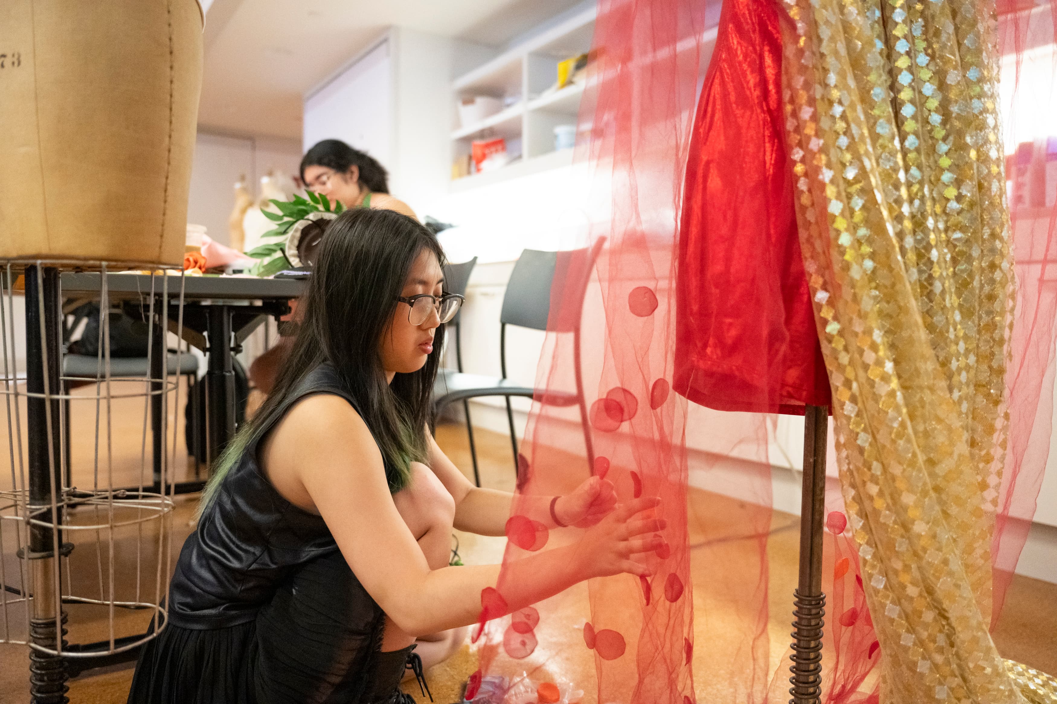 A teen crouches next to a dress form with a garment on it. The garment is a shiny red and gold dress. The teen adjusts the fabric on her design.