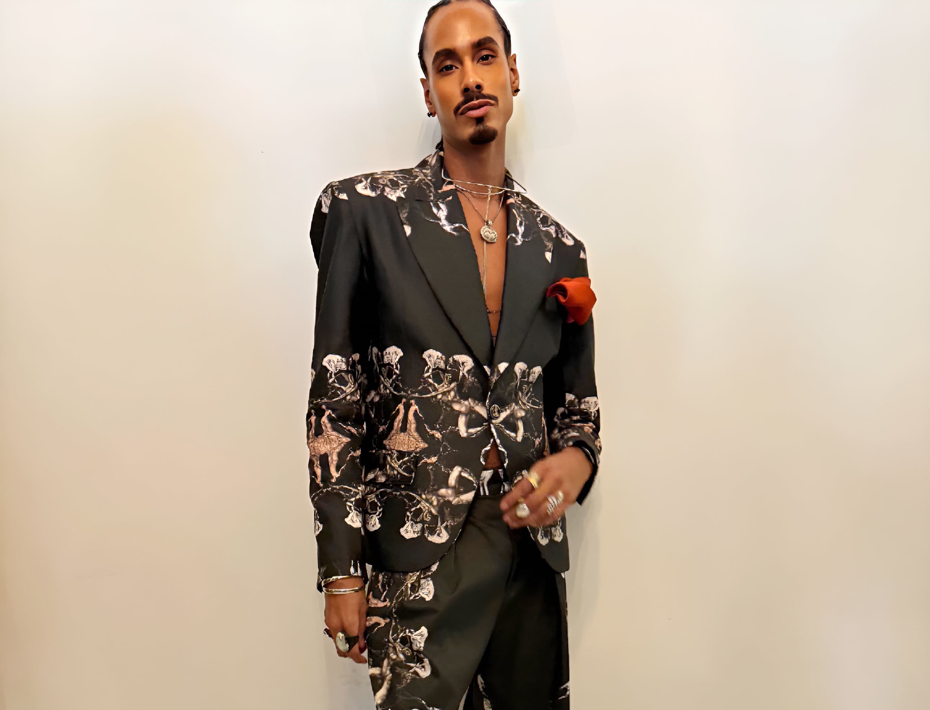 A man posing with one foot forward, wearing a black suit with light patterning and an orange pocket square.