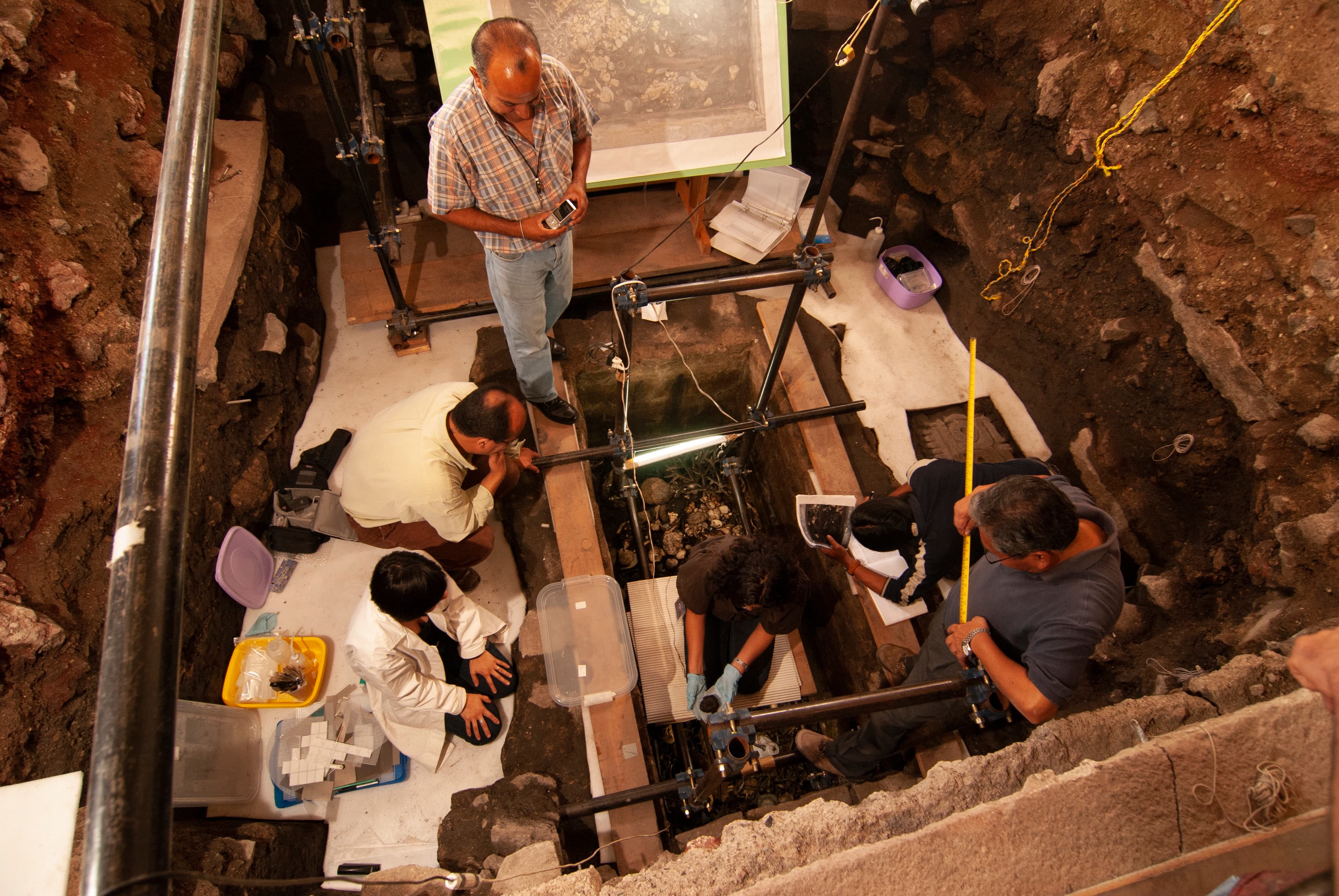 A photograph depicting an overhead view of an archaeological excavation with a rectangular shape opening in the ground and six people working on the site