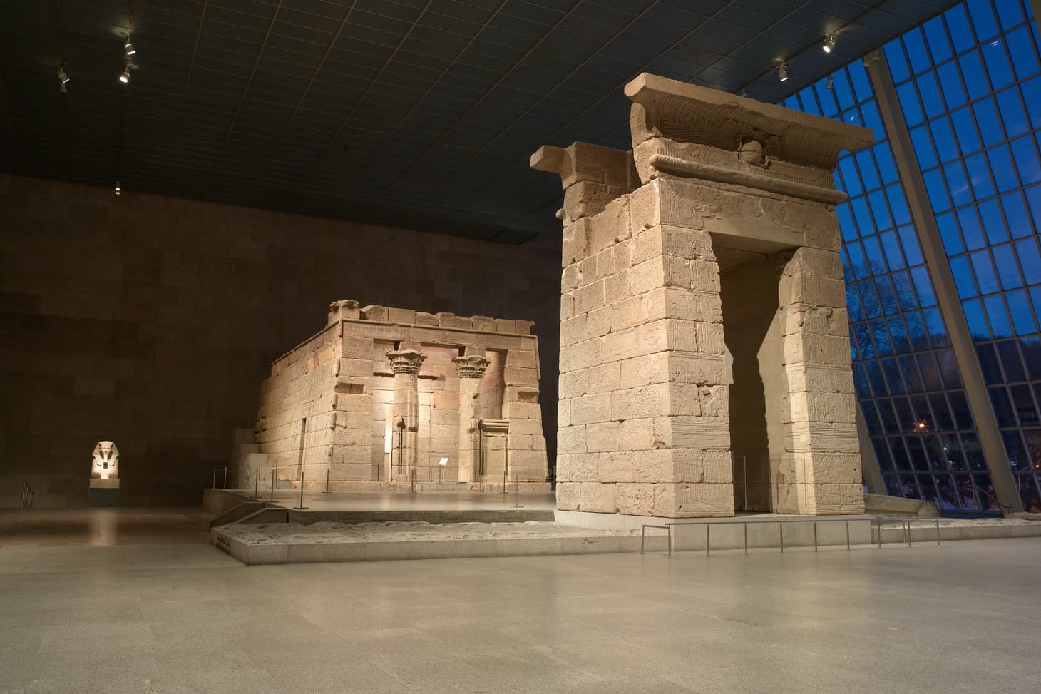 An installation view of The Temple of Dendur, a temple and gate made of Aeolian sandstone, at dusk inside of the Museum situated on a platform surrounded by water and flanked by a large wall of windows on the right overlooking Central Park.