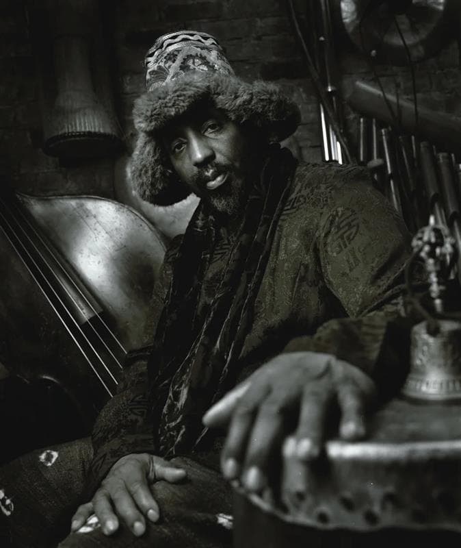 A black and white portrait of William Parker. He’s surrounded by instruments and looking seriously at the camera. The camera angle enlarges his hands in the foreground as he leans back.