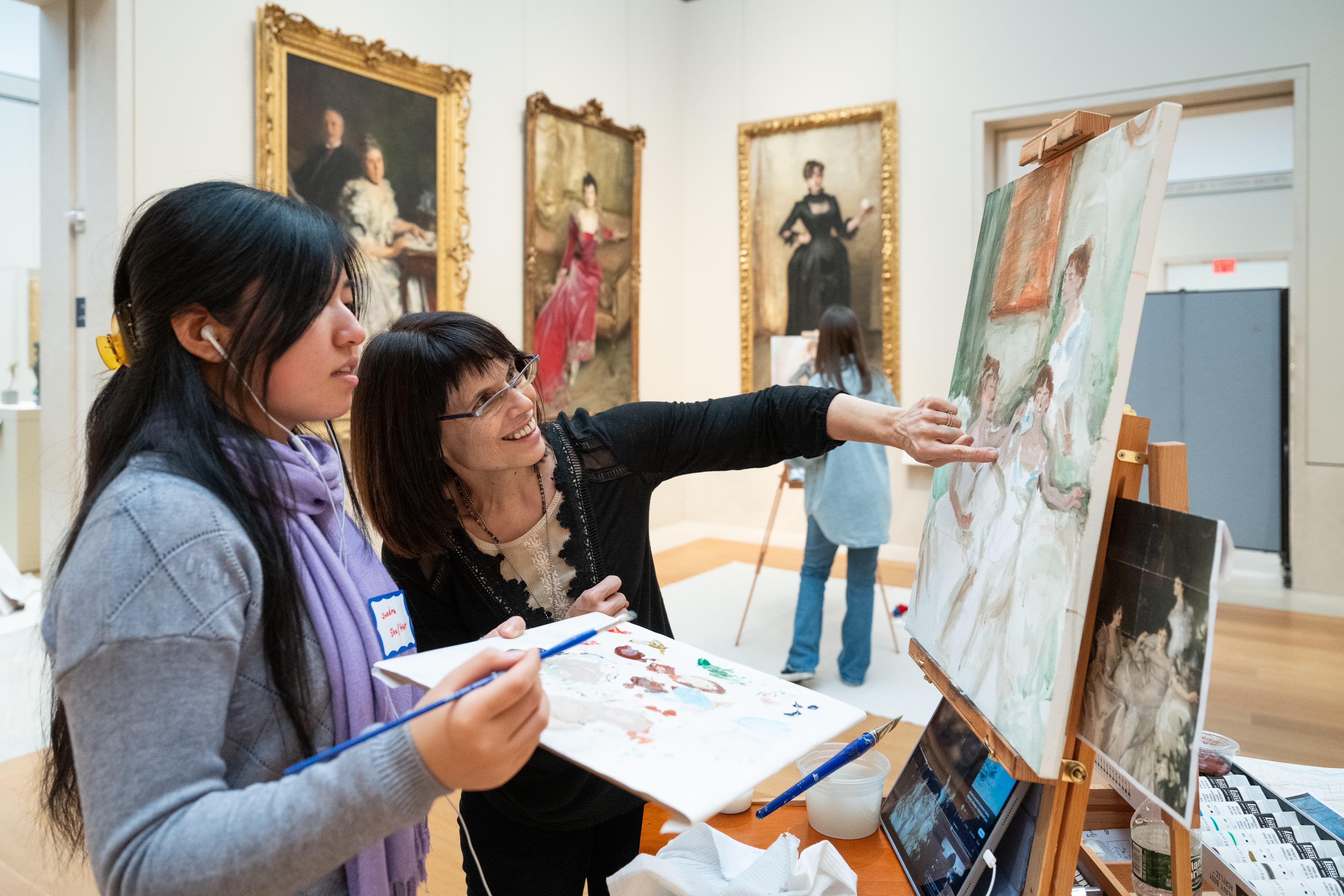 A teen artist views her in-progress painting with an educator who gestures towards her canvas. Her painting is setup on an easel and she holds a paint palate and a brush in her hand.
