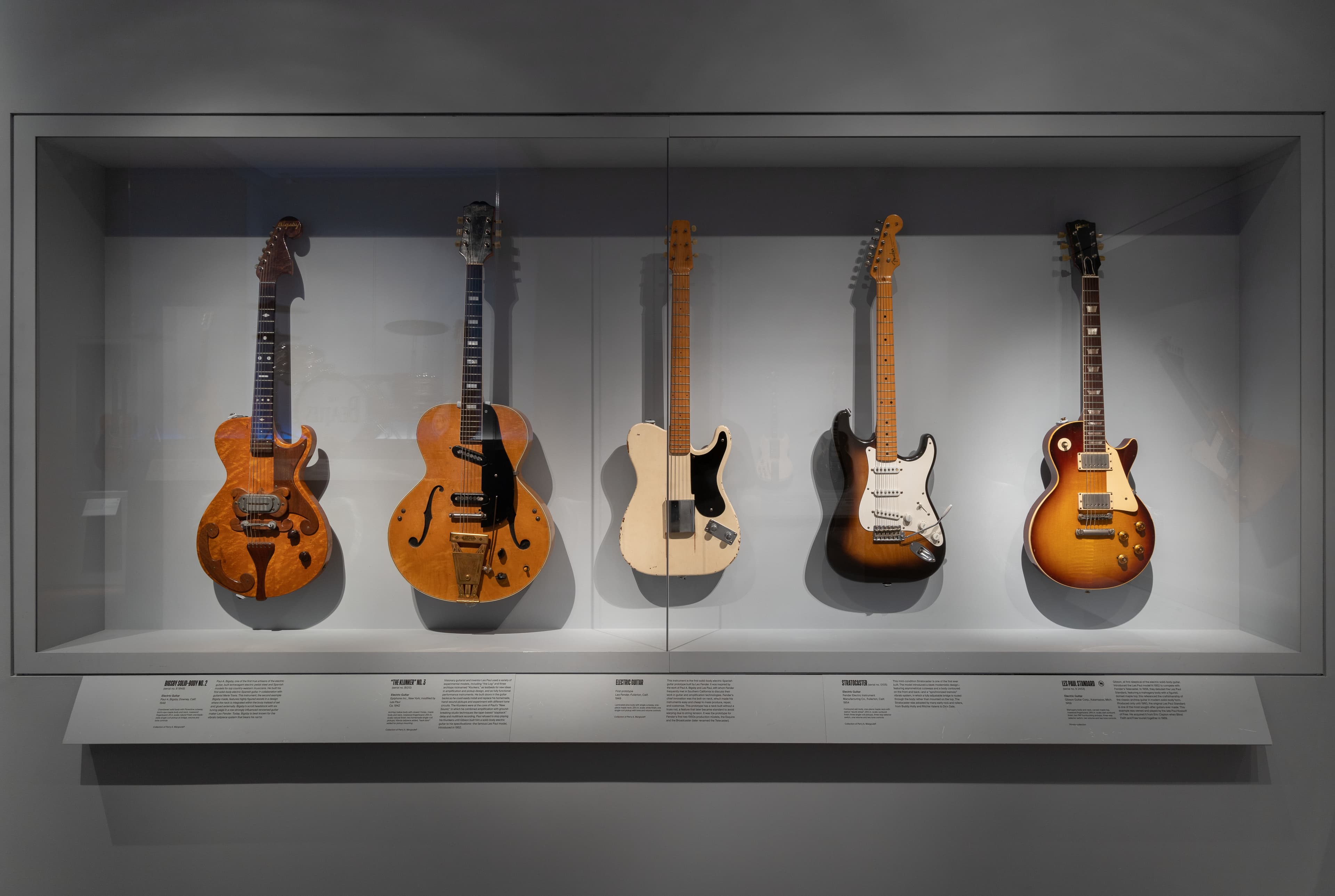 A collection of five brown and white electric guitars, displayed side by side in a display case. Photo Credit: Wilson Santiago 