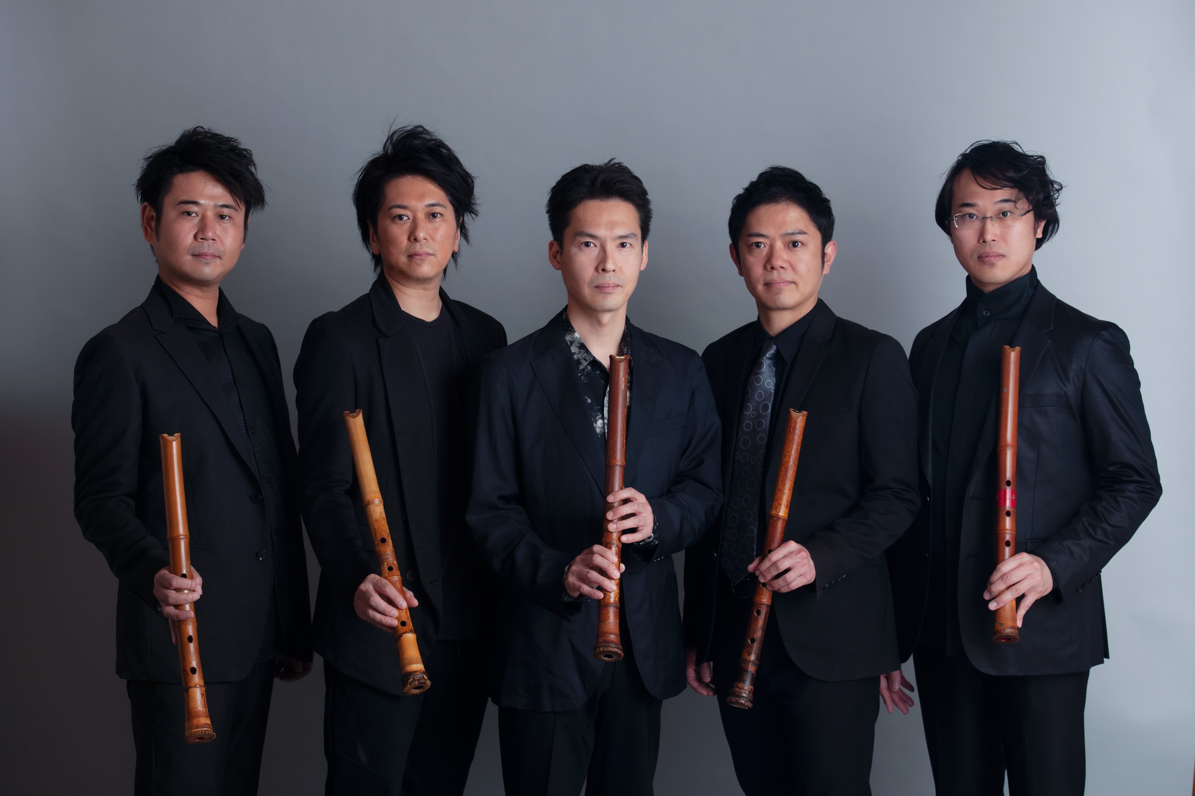 Five musicians standing together, wearing black, holding Japanese shakuhachi flutes. 