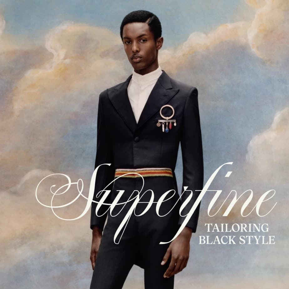 An African American man stands against a cloudy-sky background, wearing a tailored dark suit with medals and a striped belt. The text "Superfine Tailoring Black Style" is overlaid on the image.