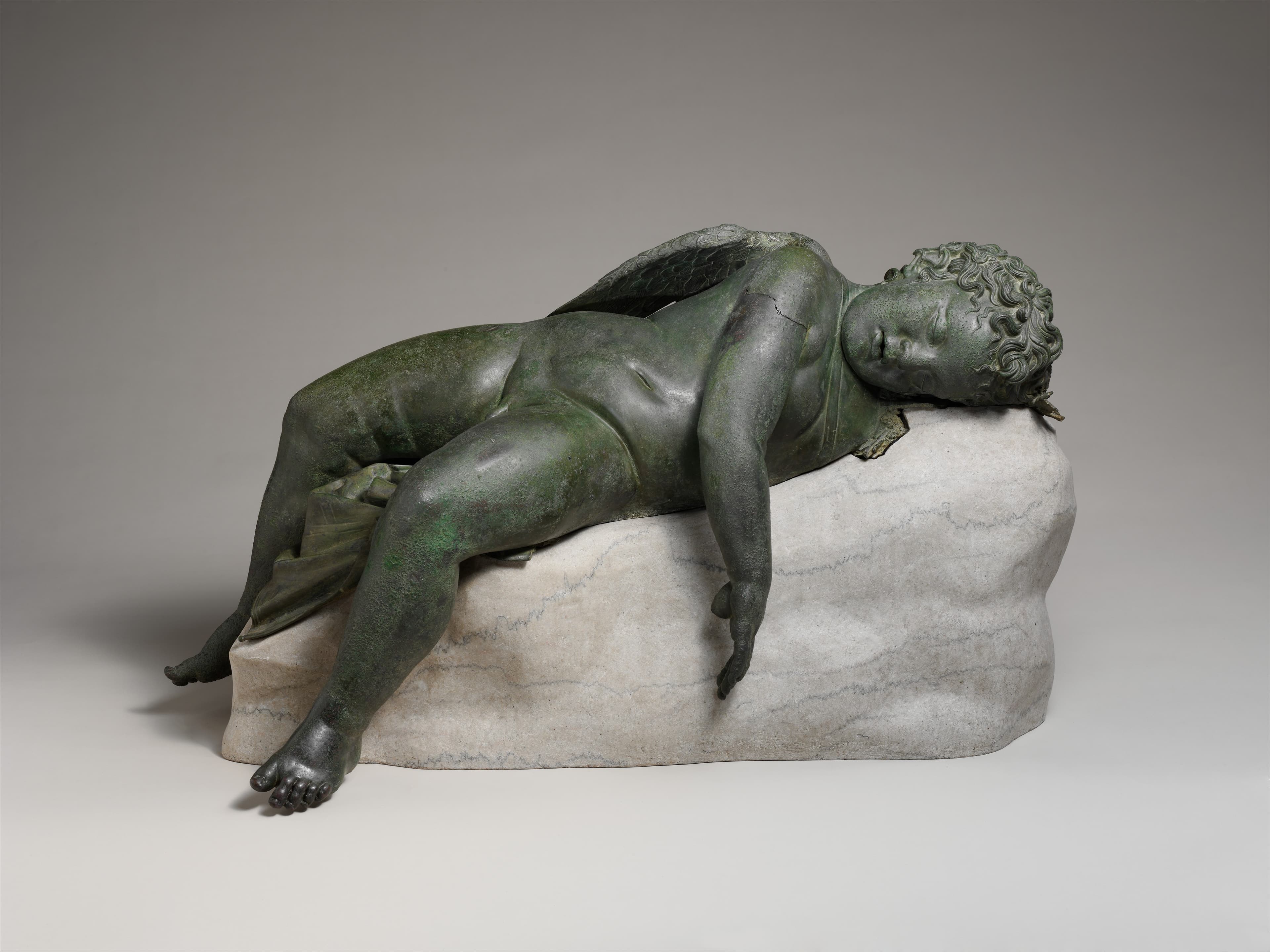 A bronze sculpture of a plump baby with wings relaxed on a stone support.