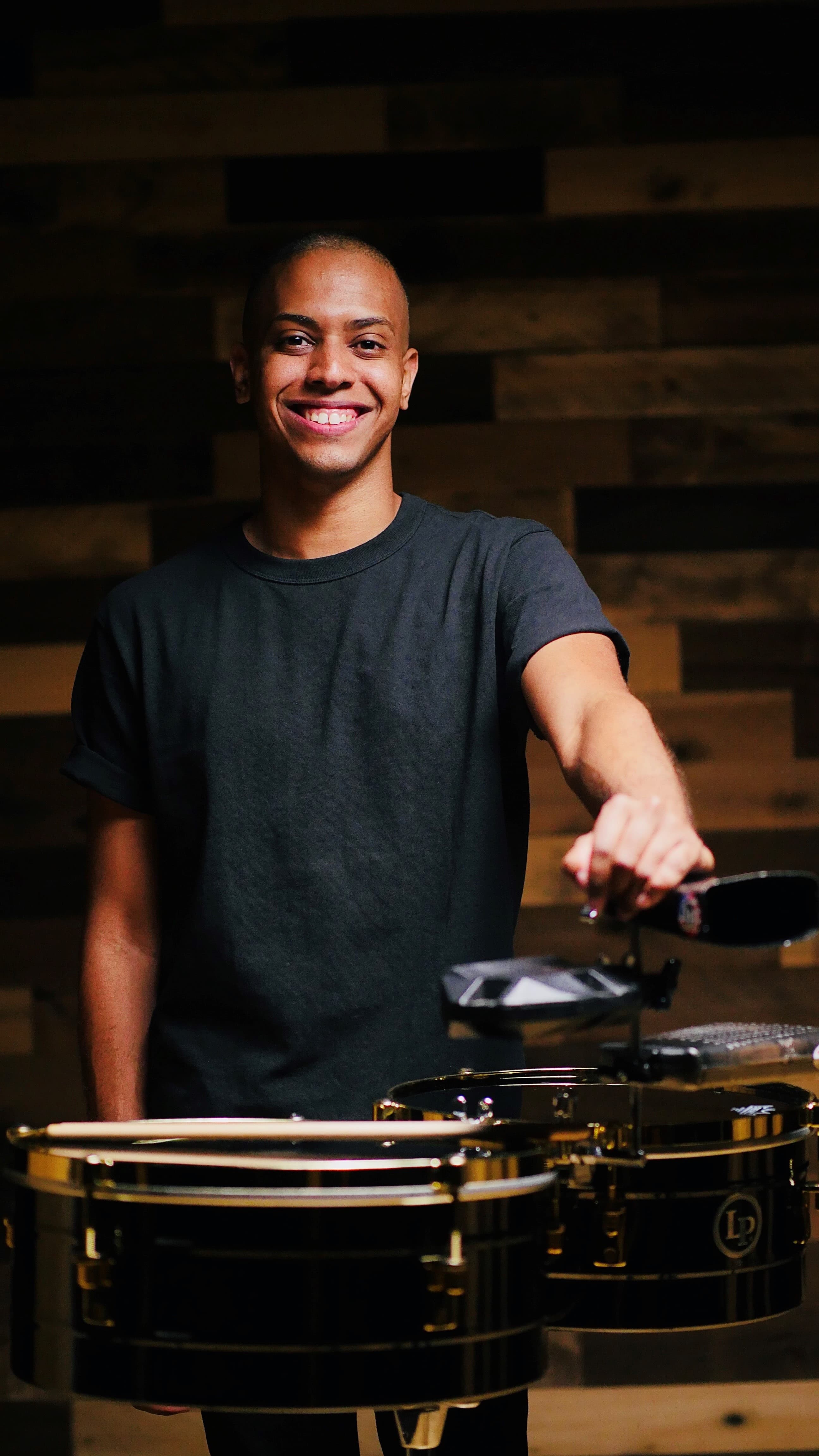 A photograph of a man standing wearing a grey t-shirt smiling as he gently leans on a set of timbales.