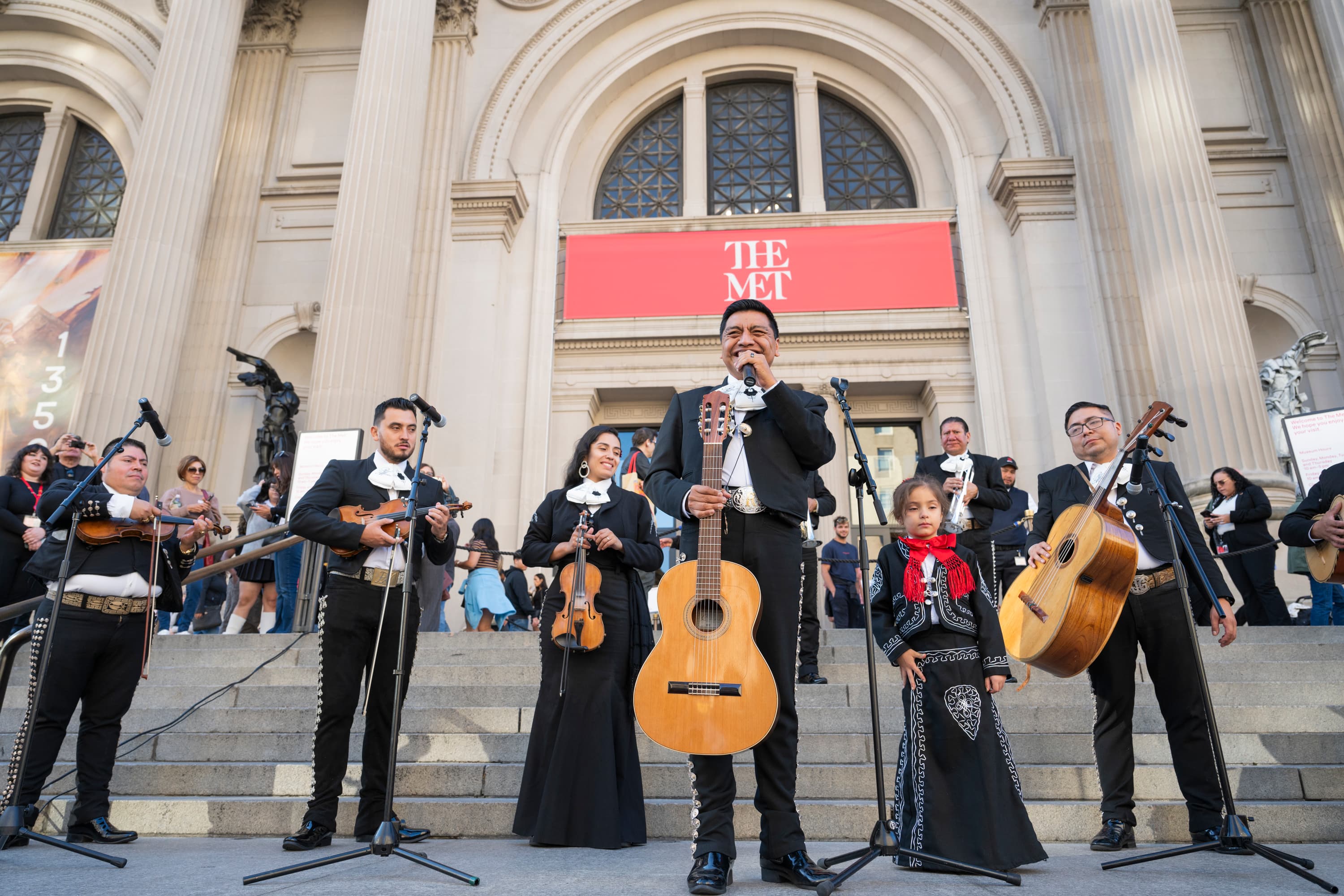 Six individuals are dressed in black, matching uniforms with tight, ornamented trousers, boots, wide bow ties, and short jackets. They are shown standing on the Museum steps, holding and performing their instruments: a violin, guitar, and a guitarrón.