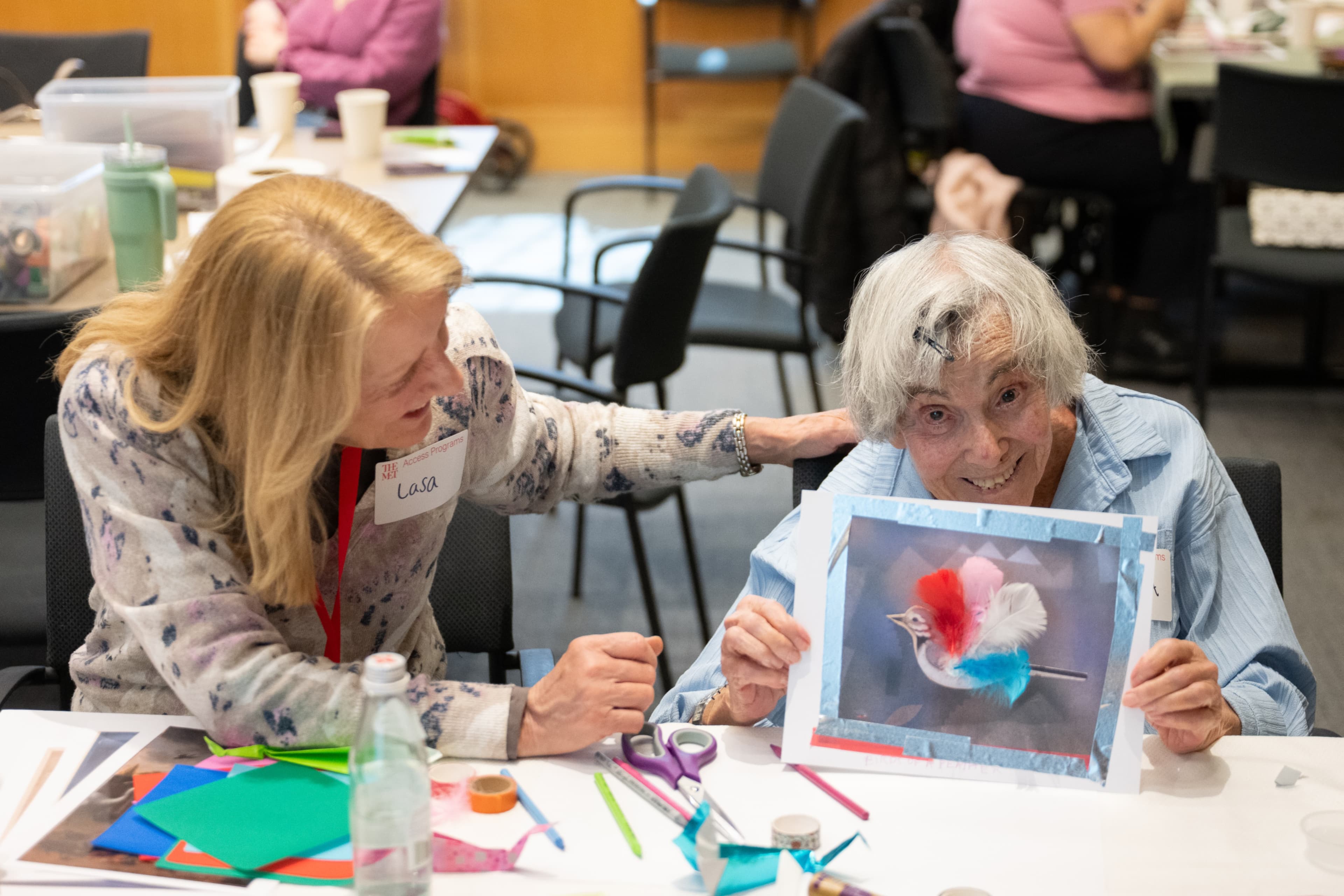 Two people sit at a table with colorful artmaking materials. The person on the left has her head tilted down and her arm and gaze extended to the woman on the right. The woman on the right looks at us with a big smile across her face while holding up a collage of a bird.