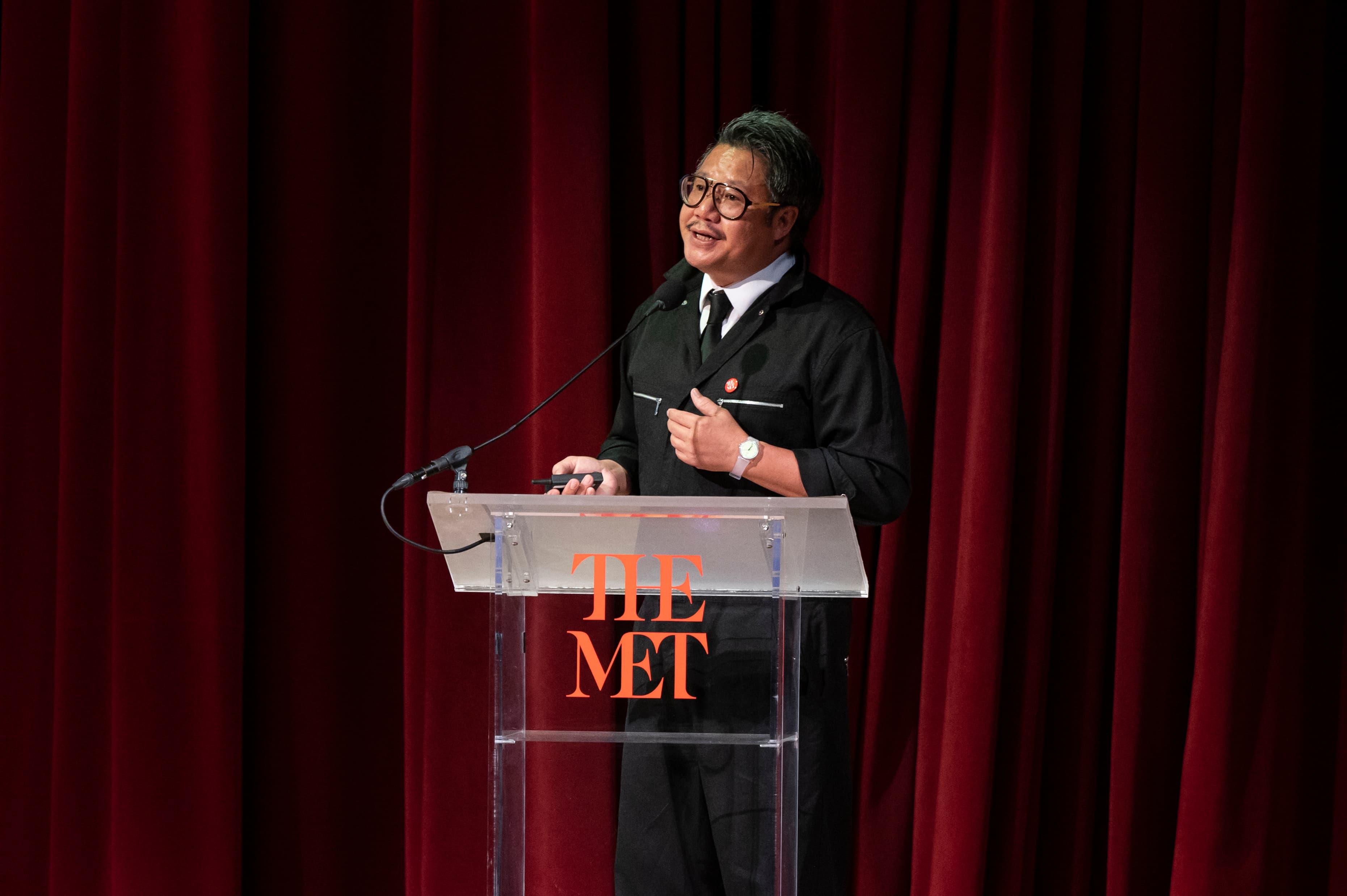 A man wearing a black jumpsuit and glasses at a lucite podium with the Met logo 