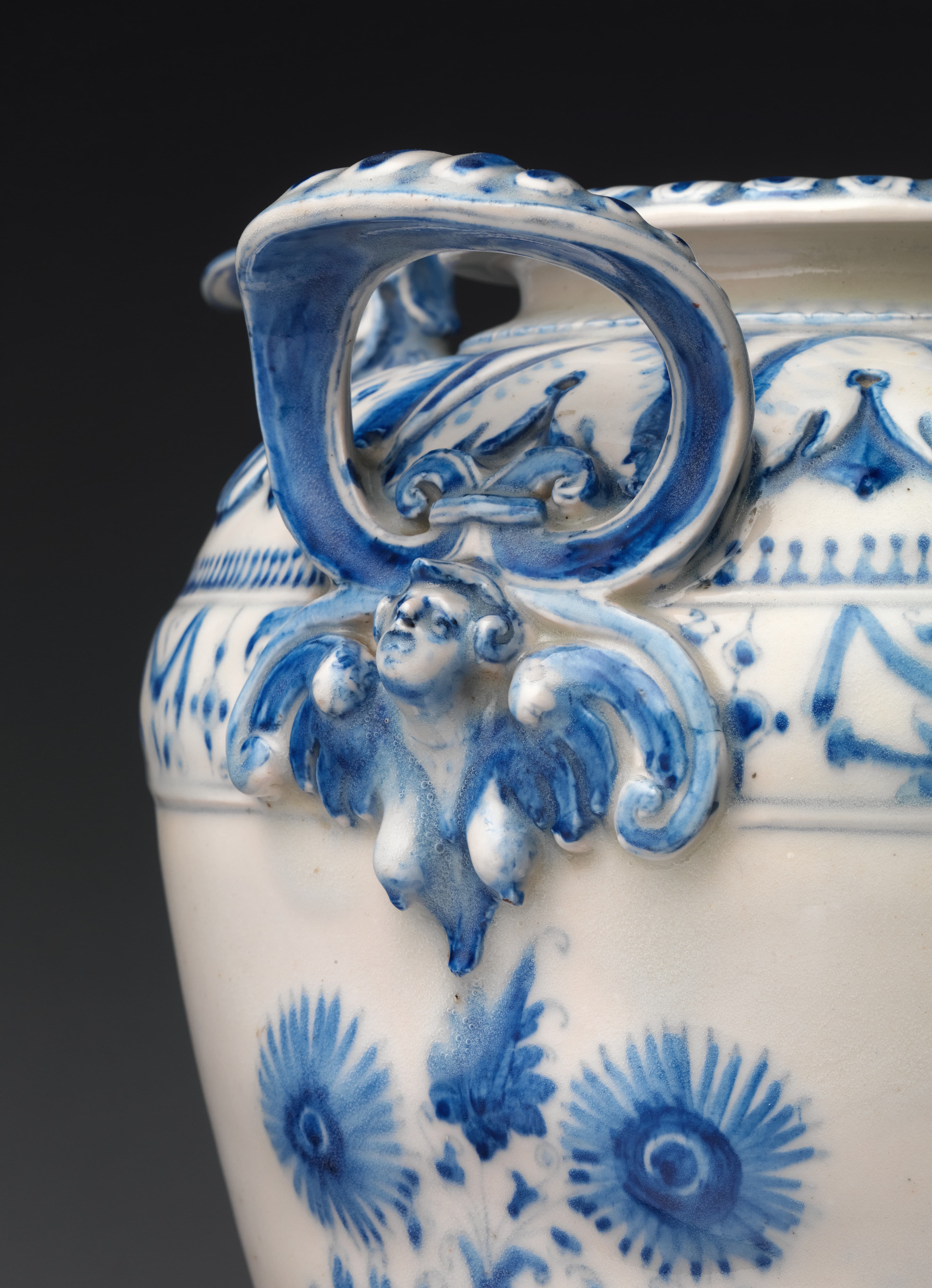 Detail of a white porcelain vase with blue decorative accents and a three dimensional relief of a face emerging from the left side.