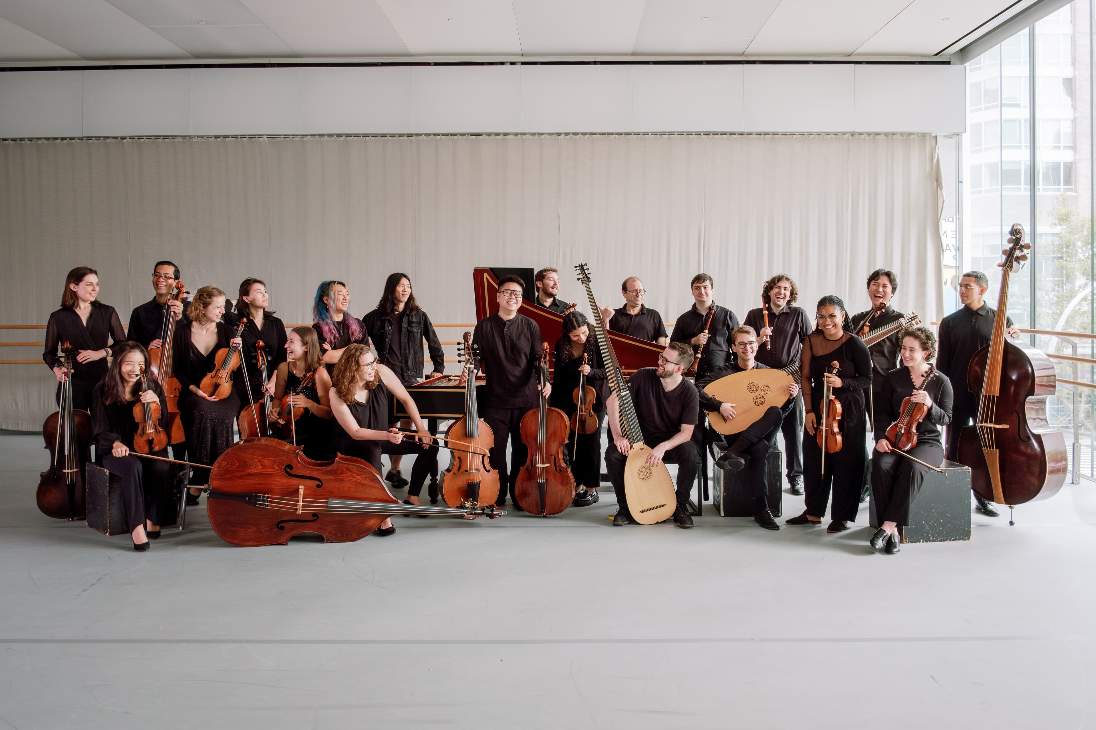 21 musicians, dressed in black, sitting and standing with their instruments, smiling at the camera and each other. 