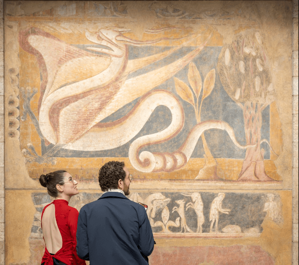 Two people stand in front of an artwork. One is wearing a red backless top with a black skirt, and the other is wearing a blue jacket and brown pants.