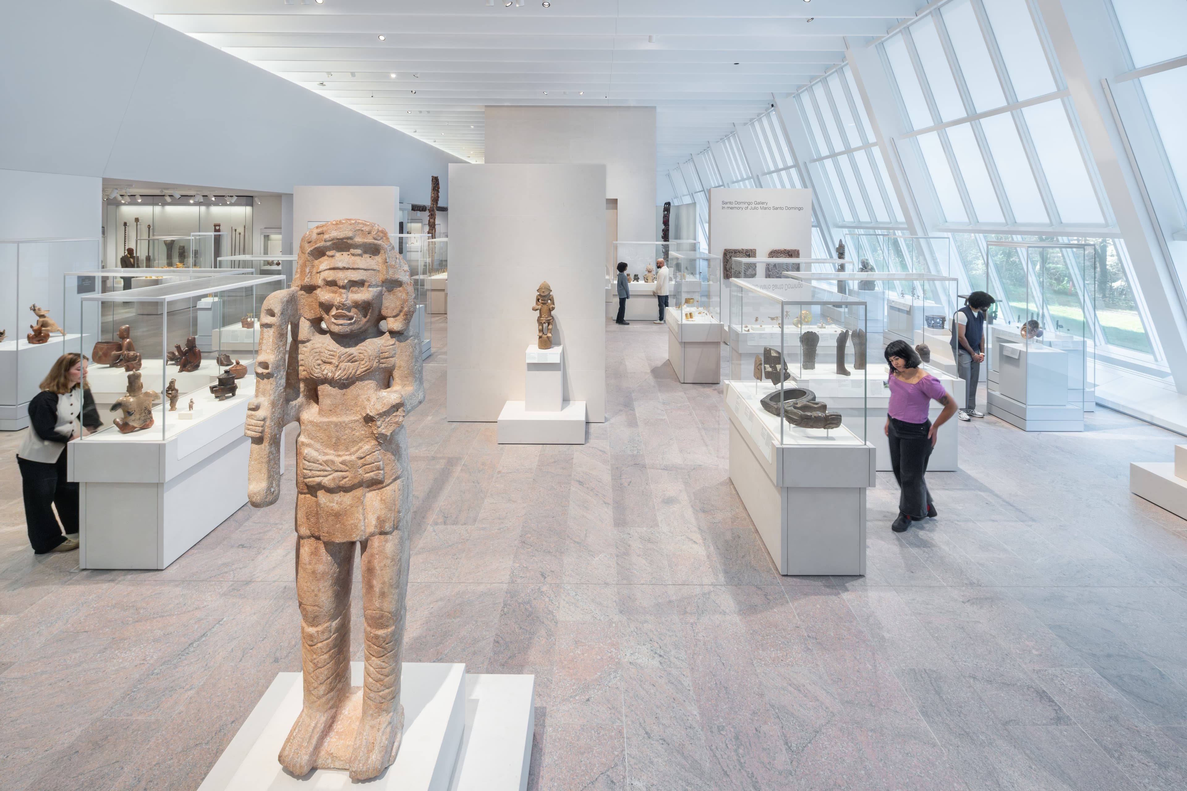 Photograph of a gallery space with floor to ceiling sloping windows on the right, cases with art objects interspersed throughout the gallery, and a large sculpture at the front left of the image. 