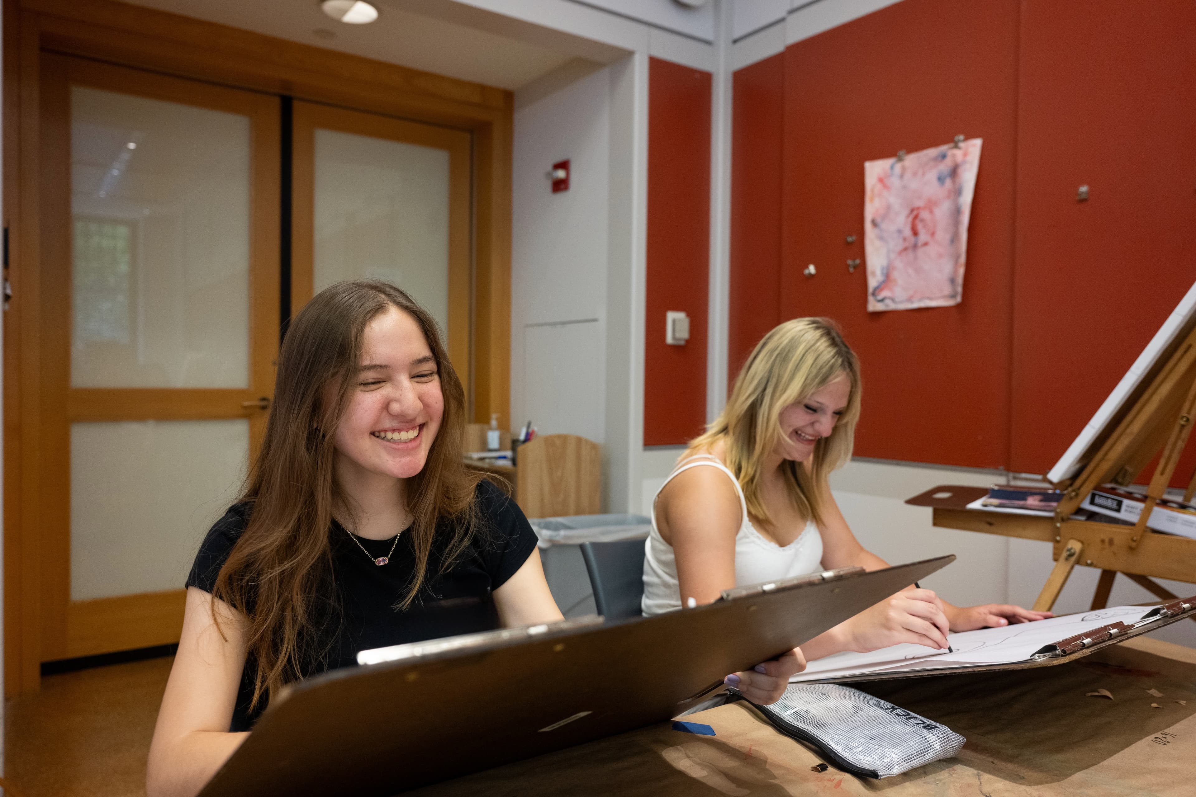 Two teens are sitting at a table in an art studio. Both are laughing while they draw on large drawing boards.