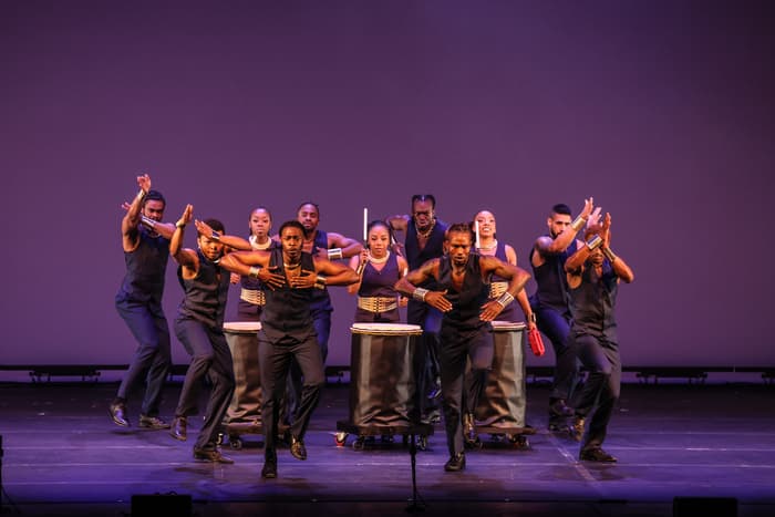 Step Afrika! during a live performance. The group of artists are looking at the camera while energetically clapping, stomping, and drumming. They are all wearing black and the stage and background are purple.