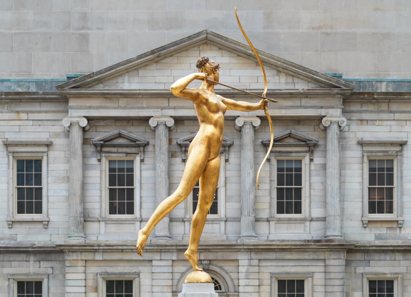  In the foreground of the image is the gilded Diana which has a gold patina, and in the background is a building with a Neoclassical facade.
