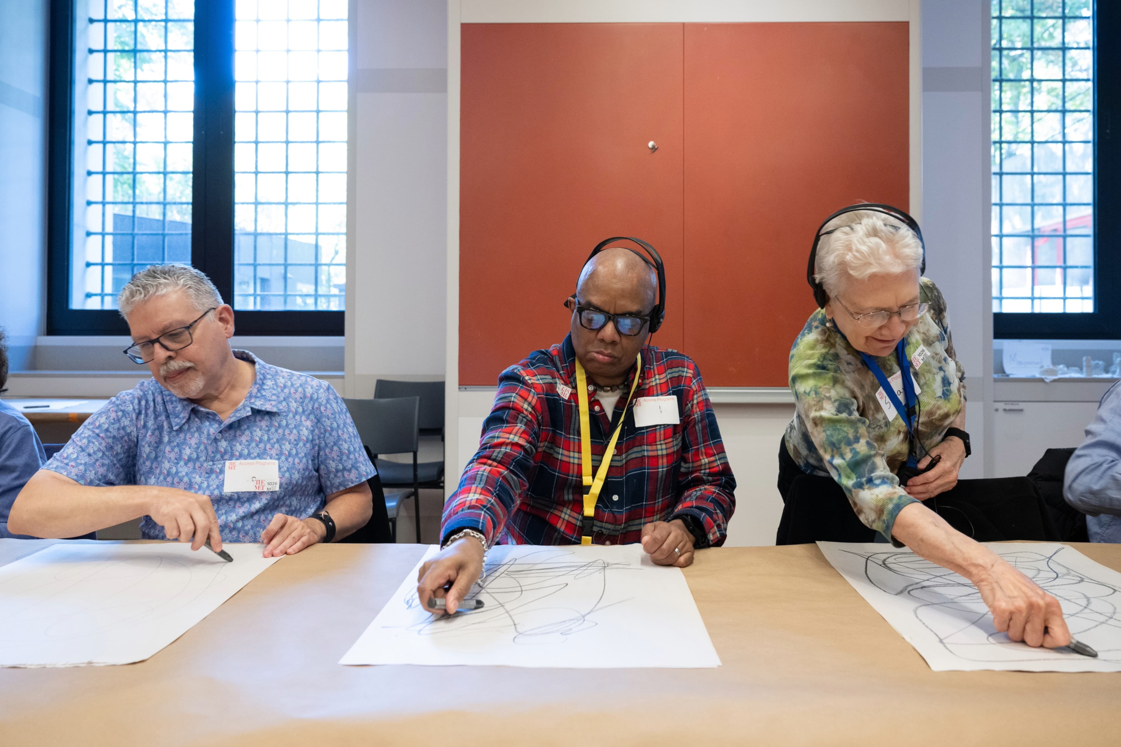 Three people draw with graphite on light grey vertical pieces of paper. A man on the left sits while wearing a blue short sleeved button up and glasses as draws at the bottom of his paper. In the middle, another man wears a dark long sleeved plaid shirt with glasses and headphones. He makes large confident marks on his paper. On the right, a woman leans over the table wearing glasses, headphones, and a green patterned top. She confidently draws at the top of her paper.