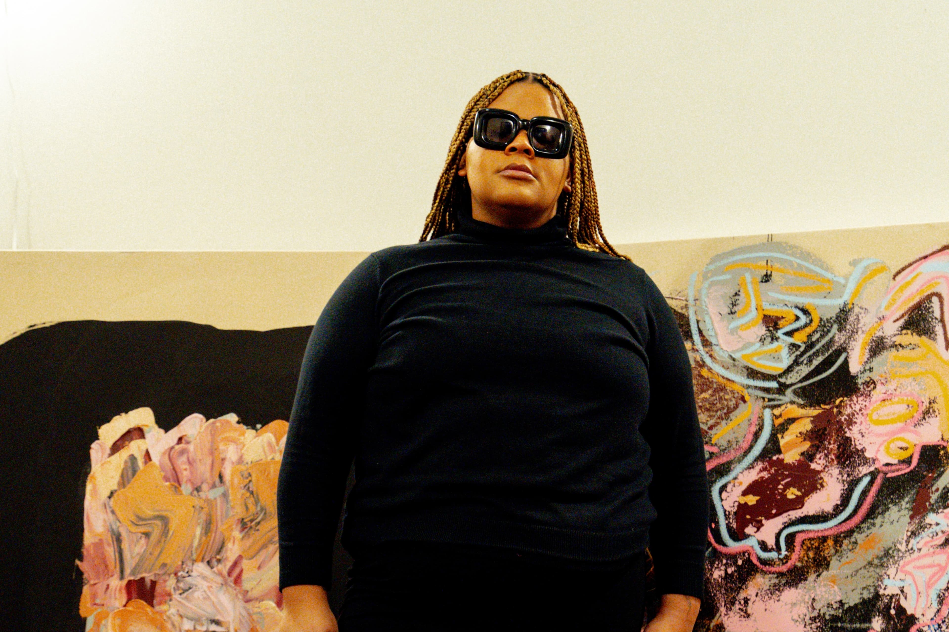 A woman stands facing the camera in front of a painting, wearing a black dress, black boots and black sunglasses.