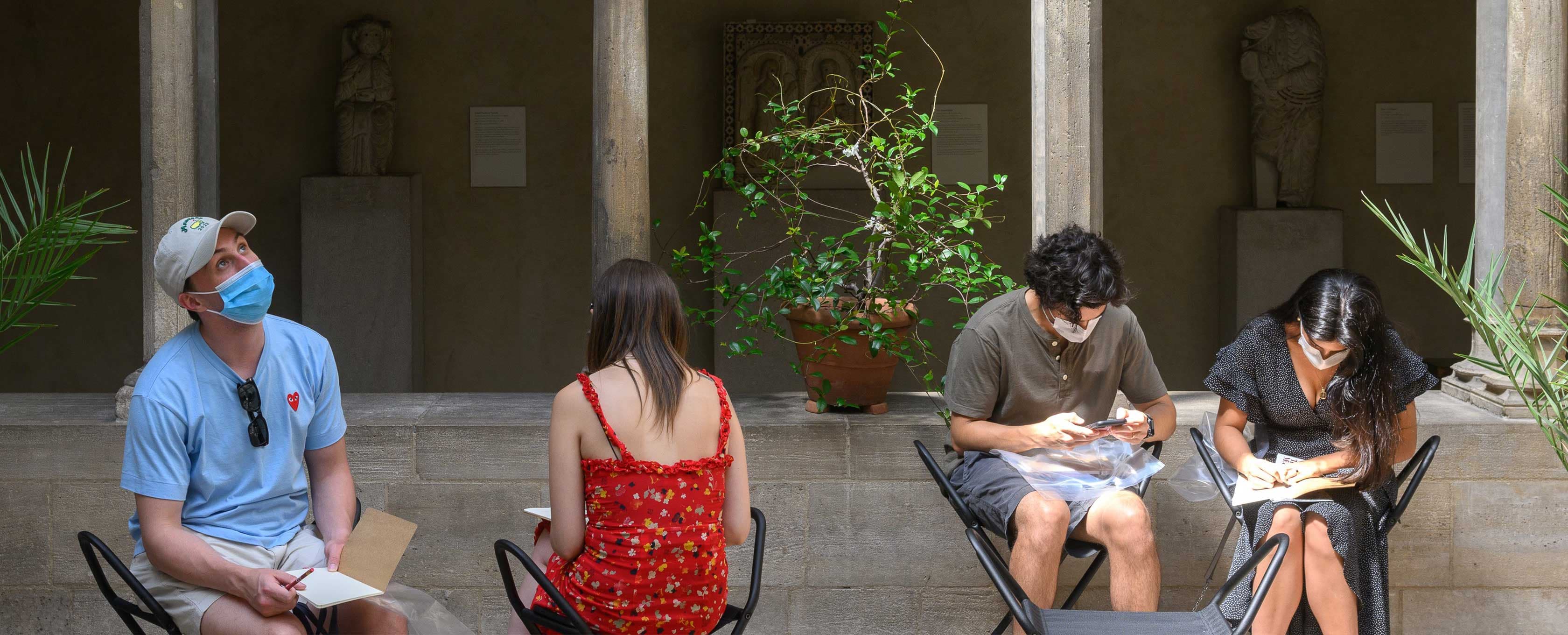 Two artists are seated on stools in an open courtyard with sunlight streaming in, surrounded by columns supporting rounded archways. There are three potted plants. Two artists lean forward over their sketchbooks, one artist looks upwards, and a fourth is facing with her back towards the viewer.