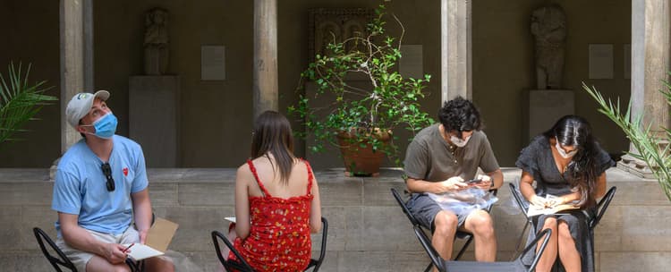 Two artists are seated on stools in an open courtyard with sunlight streaming in, surrounded by columns supporting rounded archways. There are three potted plants. Two artists lean forward over their sketchbooks, one artist looks upwards, and a fourth is facing with her back towards the viewer.