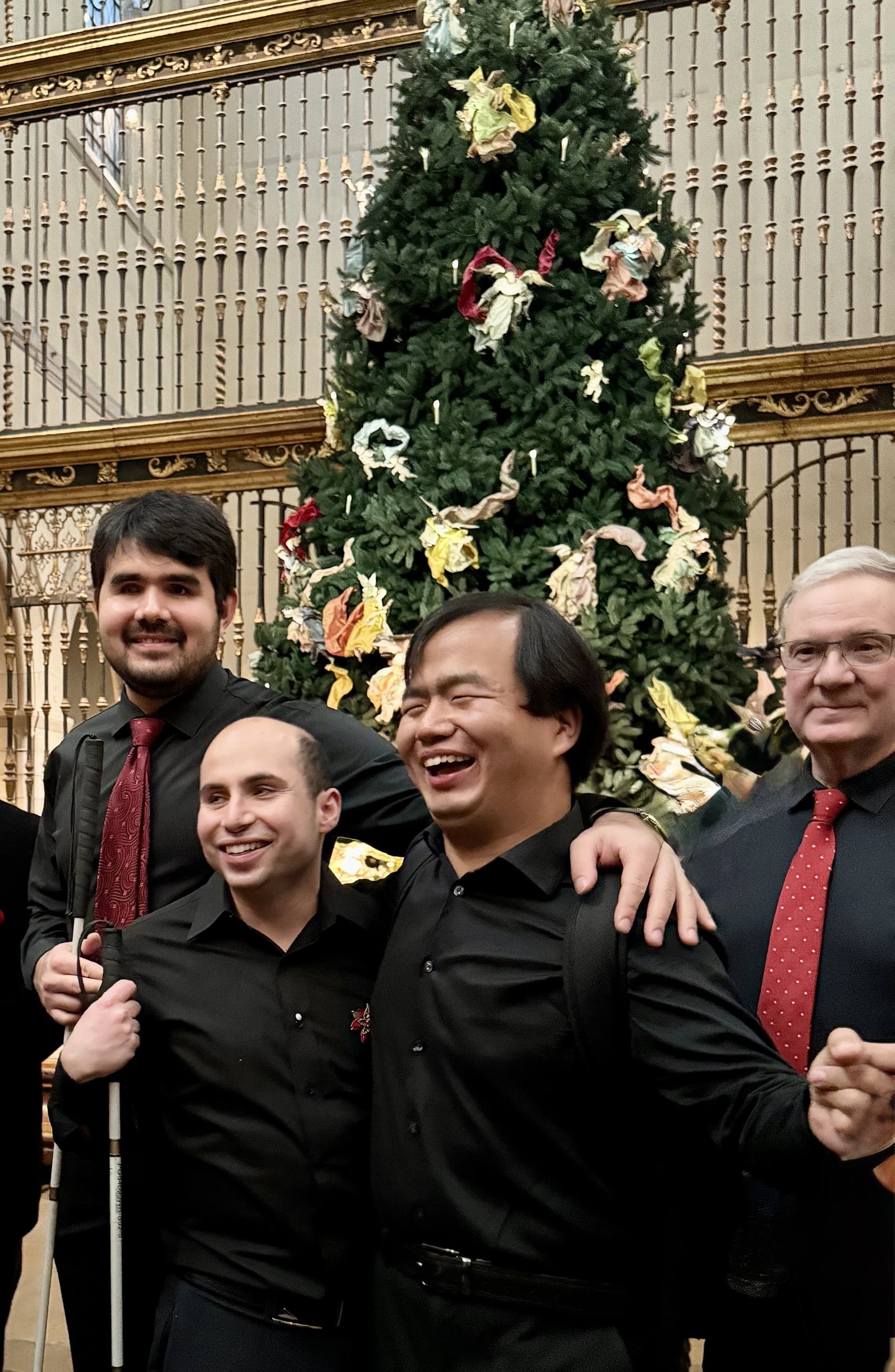 A group of four performers stand smiling in front of a Christmas tree. 