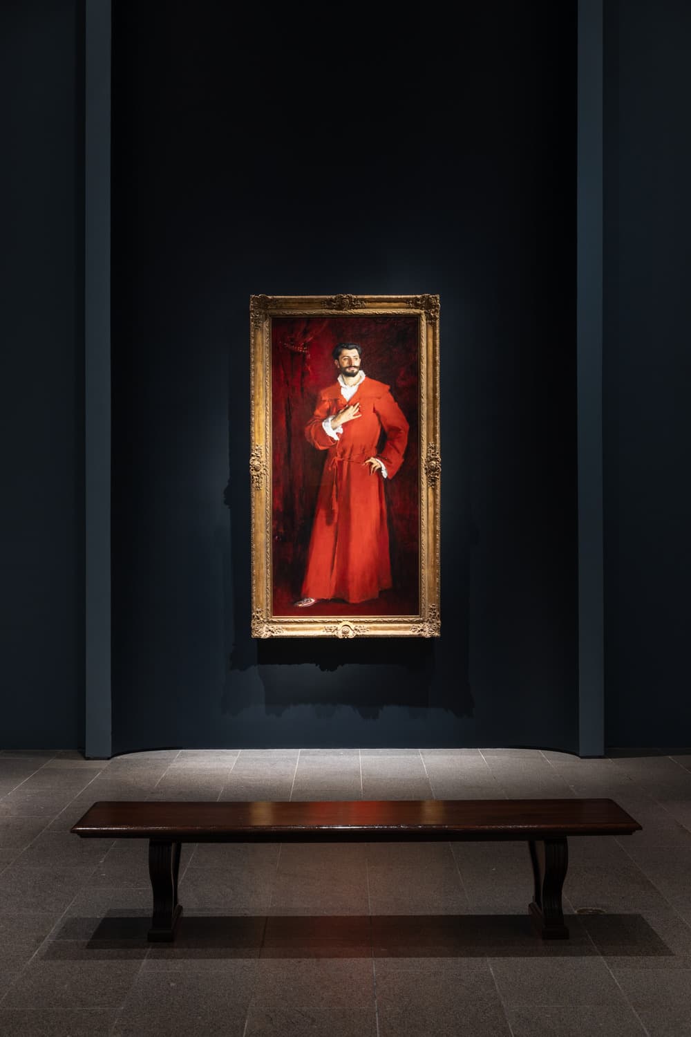 A man wearing a red velvet robe in front of red drapery stands posing with his gaze looking to the side with fingers delicately adjusting his collar and belt.