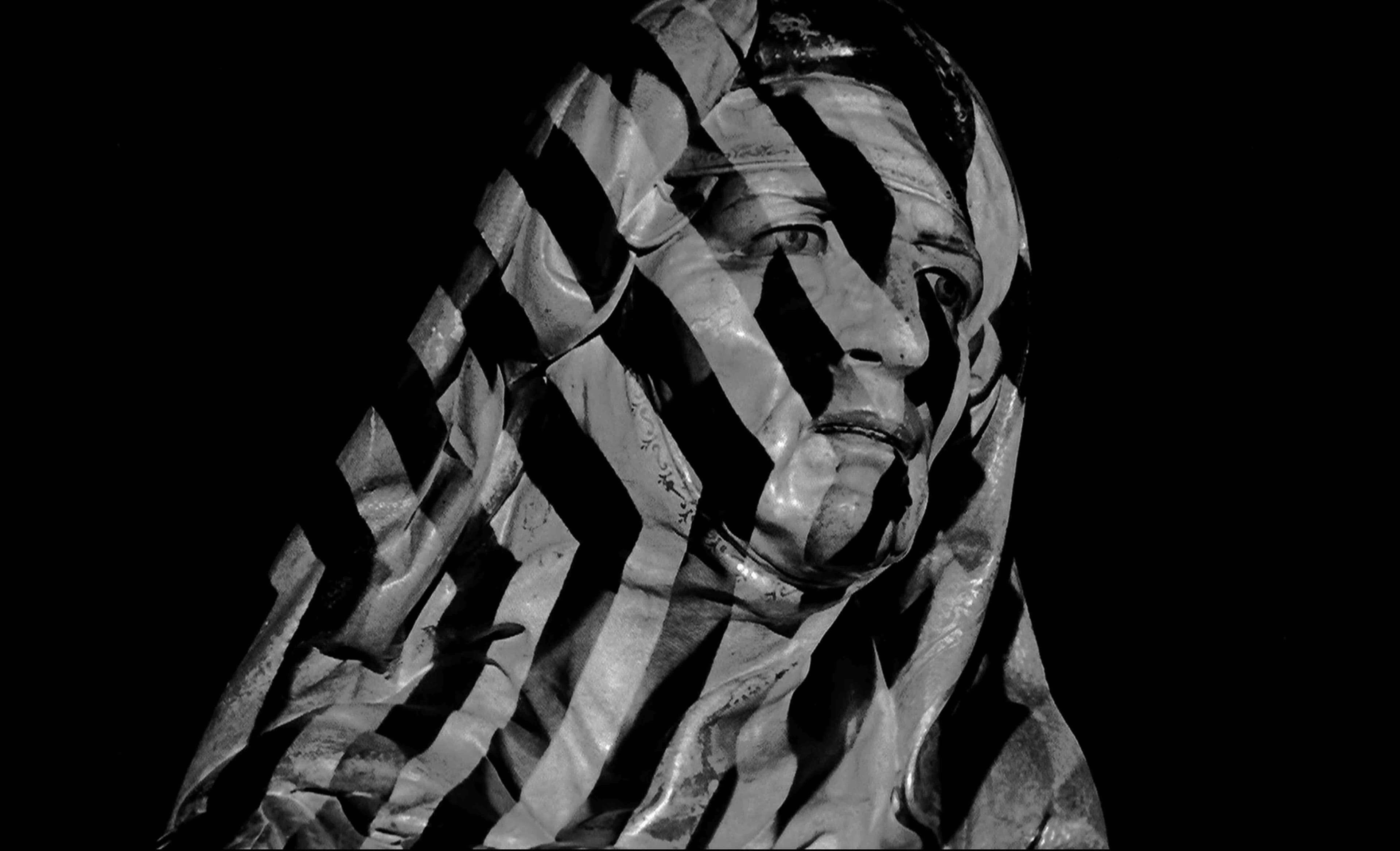 A black and white still from a film shows a sculpture of a person wrapped in stripes and wearing a head scarf.