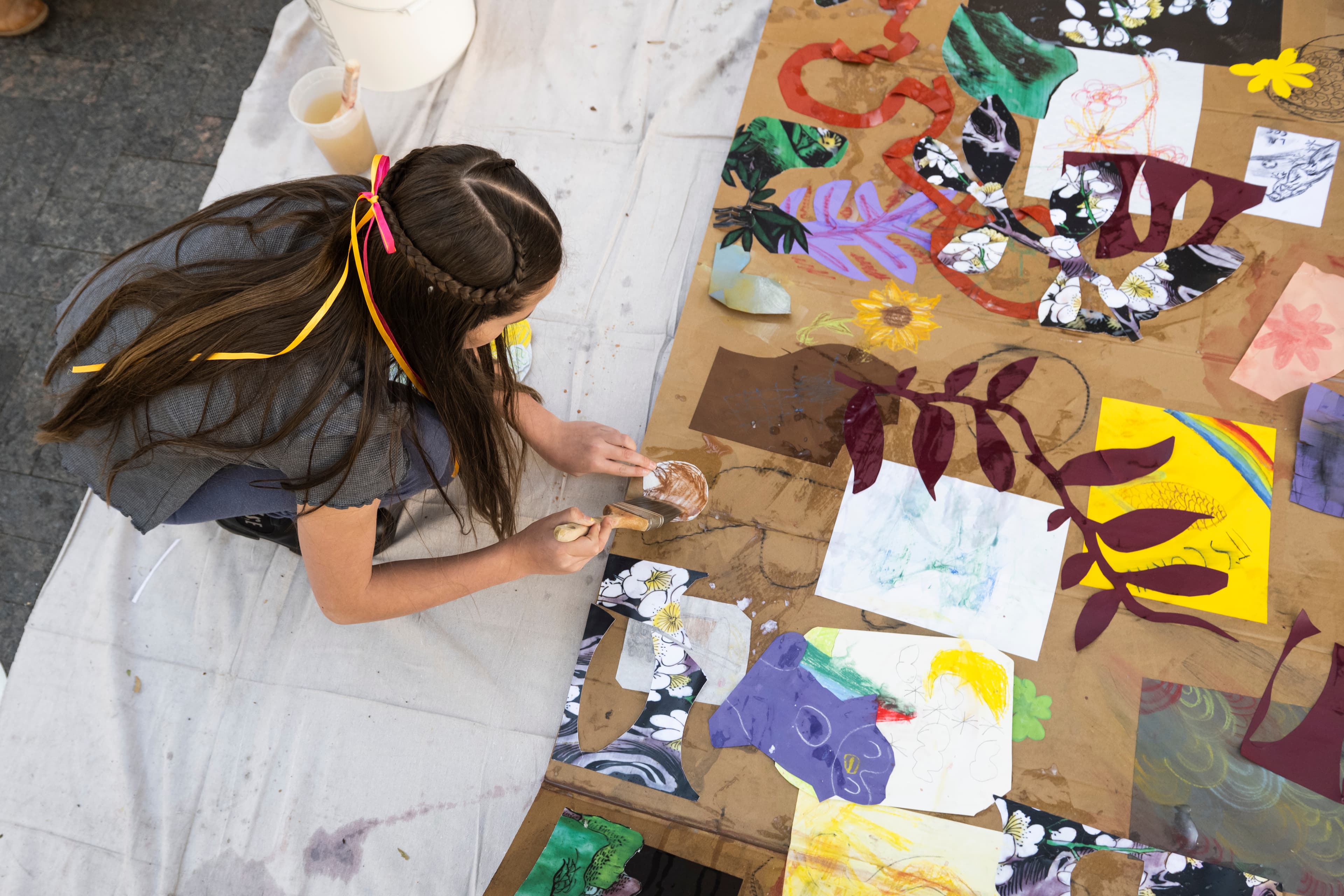 A young girl sits and paints on a large canvas. The girl is wearing dark clothes and a pink and yellow ribbon in her hair. The canvas has shapes and flowers in various colors and materials.