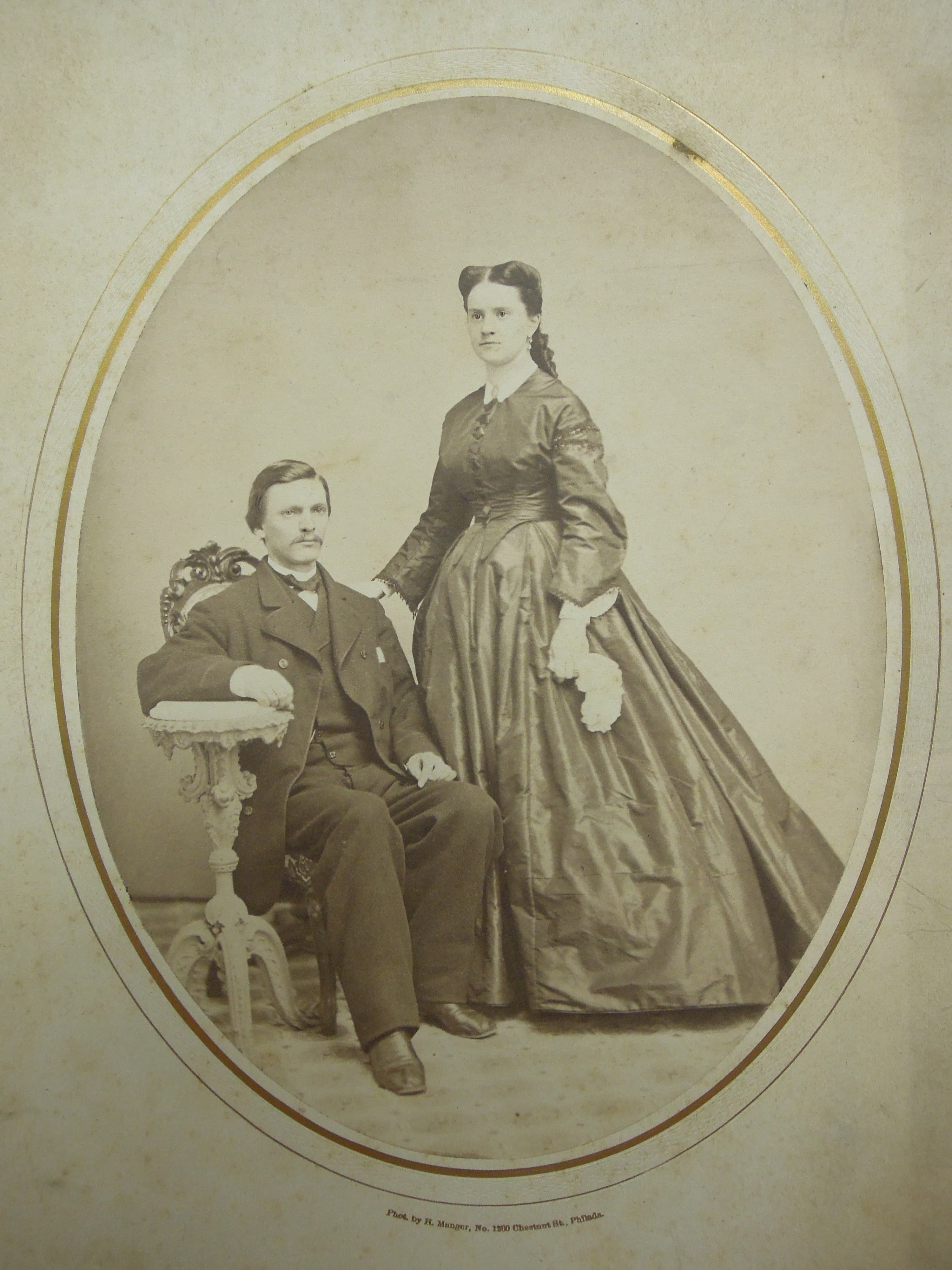 Washington Roebling and wife, Emily Warren Roebling in a black and white portrait. Washington is seated in a chair and wearing a black suit, and Emily stands over him in a dark long dress.  