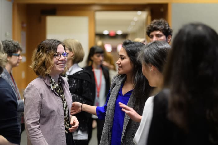 A group of people engage in conversation in a hallway. One woman in a mauve top, animal print scarf, and glasses is smiling while talking to another woman in a blue top and gray vest. Others are conversing in the background.