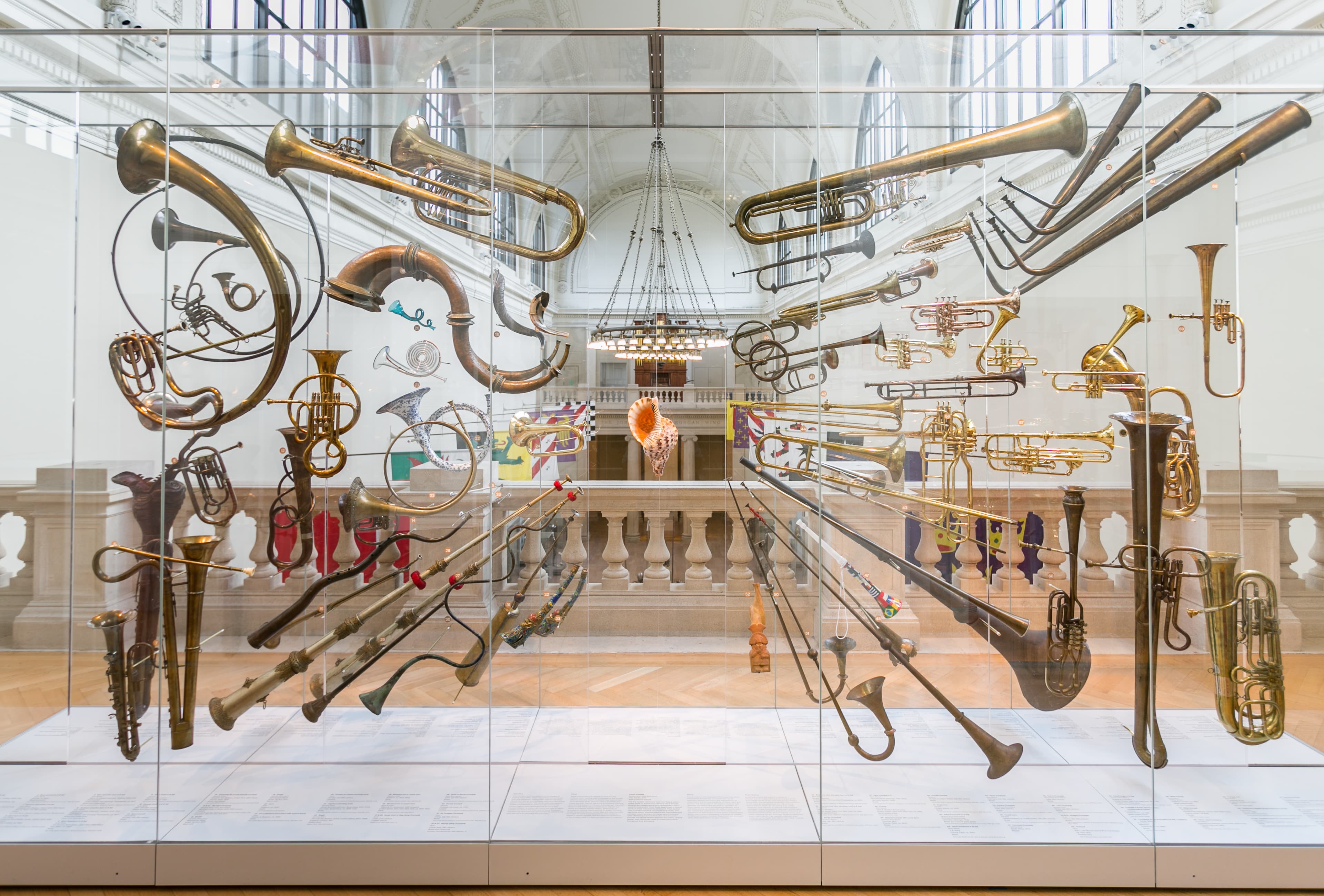 A collection of brass wind instruments including trumpets, saxophones, and trombones hang suspended in various poses in a glass display cabinet in front of an ornate, stone balcony. The centerpiece of the display is a suspended conch. 