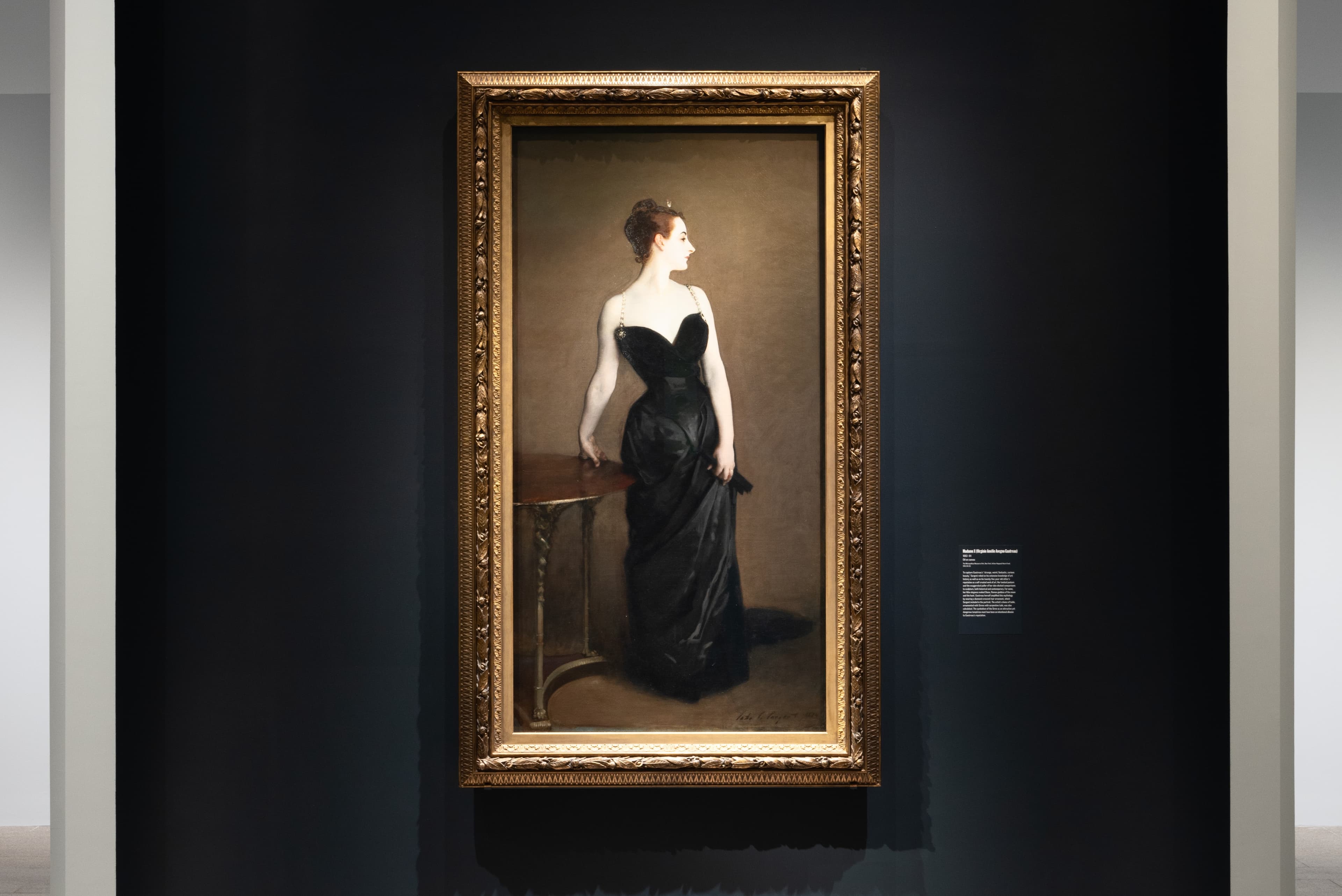 An installation image of Madame X, a woman in a black dress looking over her shoulder, on a blue wall