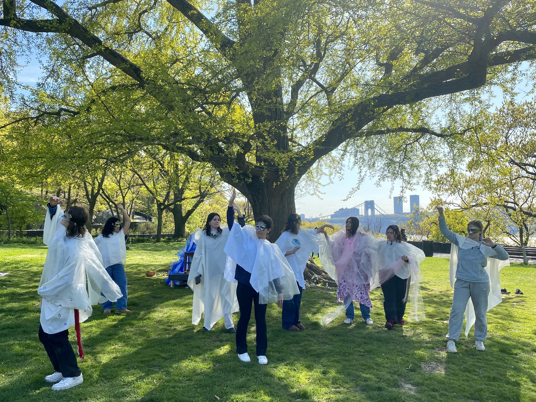 Masked performers stand underneath a tree with their arms stretching towards the sky.