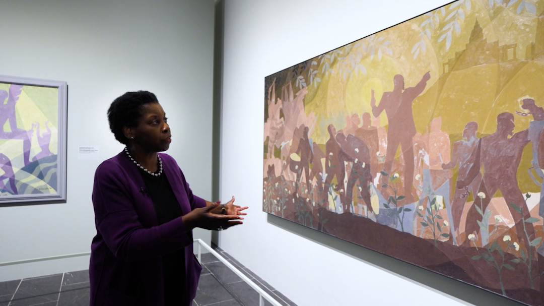 Curator Denise Murrell stands in a purple shirt before a painting by Aaron Douglas featuring geometric abstracted figures rendered in a wide range of purples and greens.