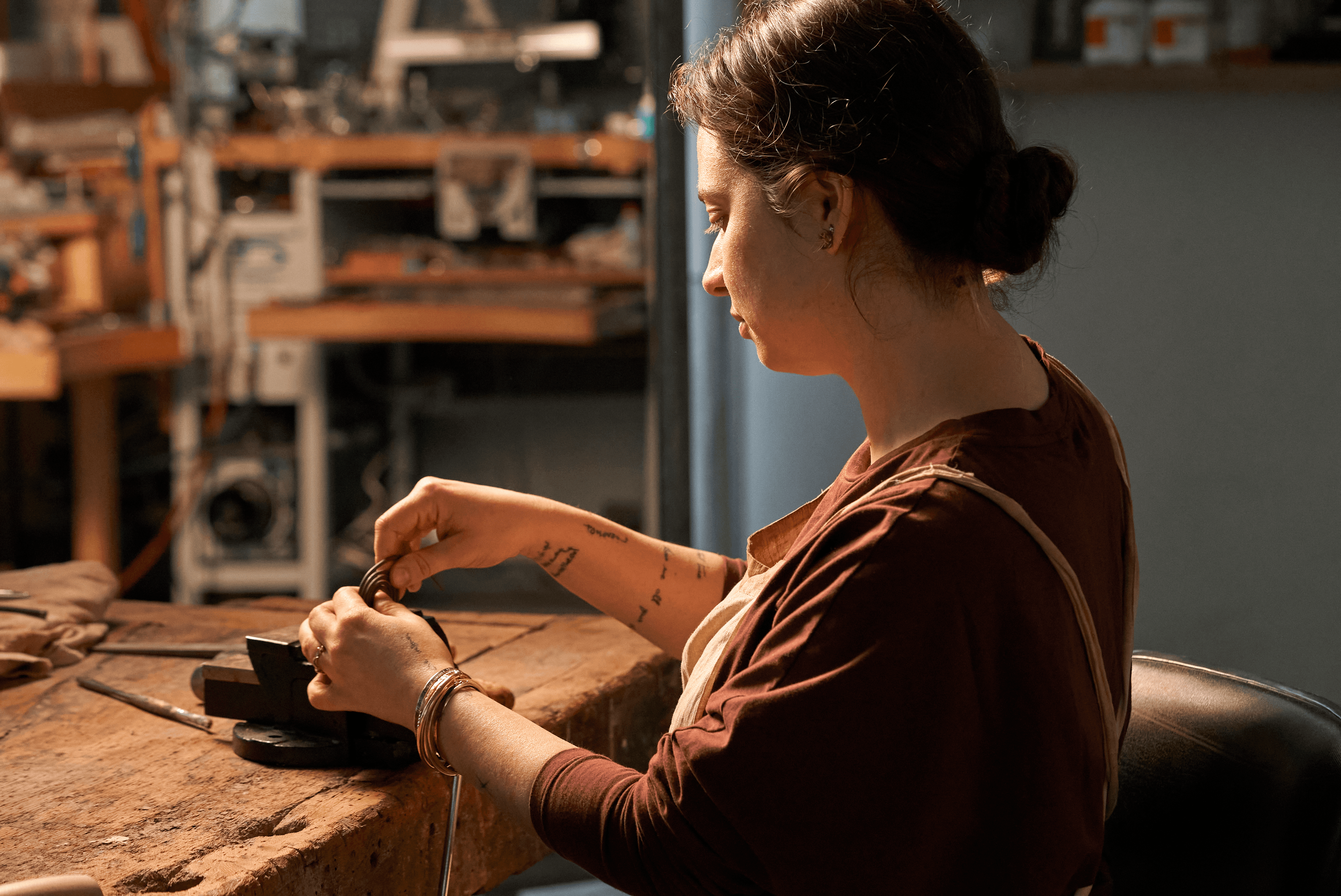 A woman artist in a jewelry studio uses a small tool at a workbench, with shelves of tools behind her.