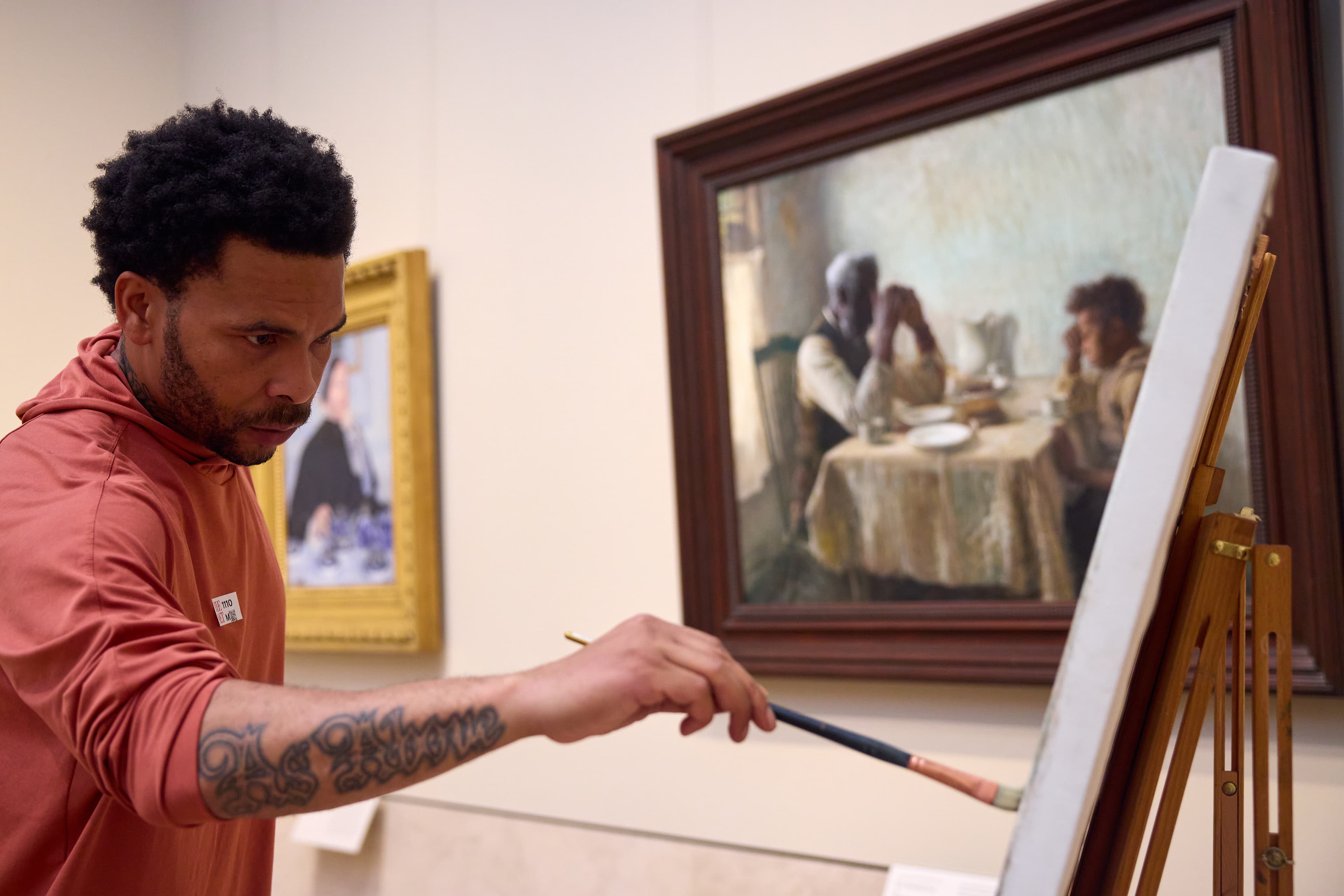 A Black man with short hair wearing an orange sweater is seen in profile painting on a canvas in a museum gallery in front of Henry Ossawa Tanner’s painting The Thankful Poor which depicts two Black people—a grey-haired man and a young child—as they pray over a dinner table.