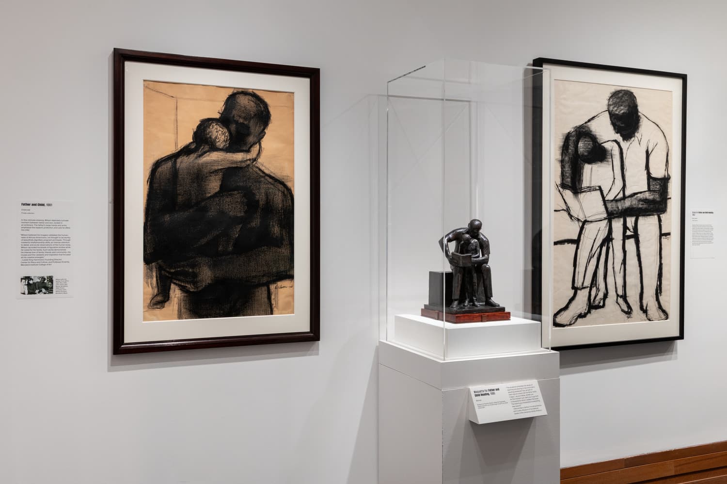 An installation view of a drawing, sculpture, and print of a man embracing a child on his lap while reading a book in front of them.