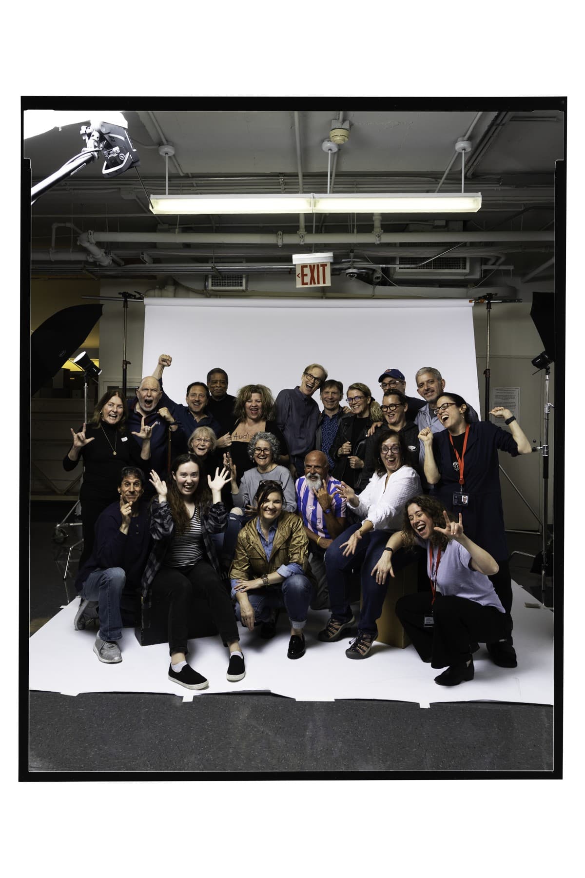 A large group of people pose together – some are signing “I love you” or are cheering in ASL. The group is on a large white backdrop with lighting and camera equipment in the background.