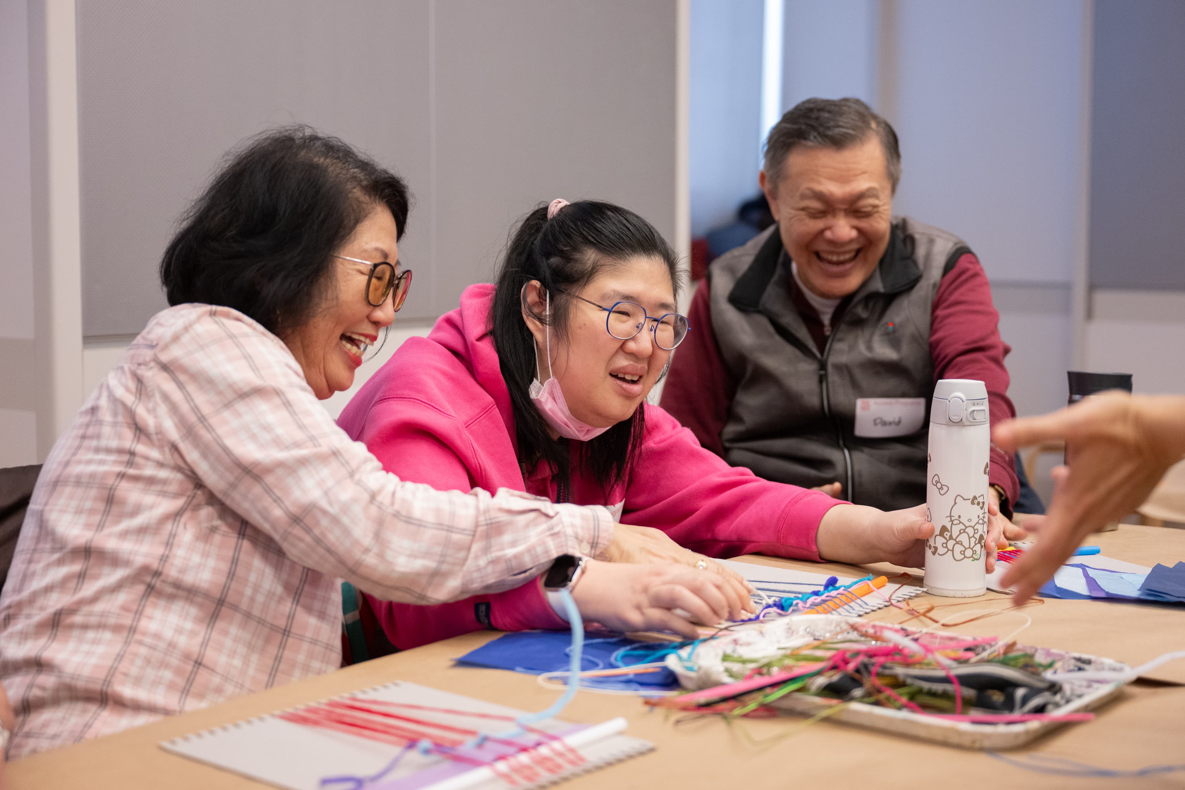 Three visitors sit at a table in a studio classroom at The Met as they work on a weaving project with yarn, pipe cleaners, and cardboard looms.