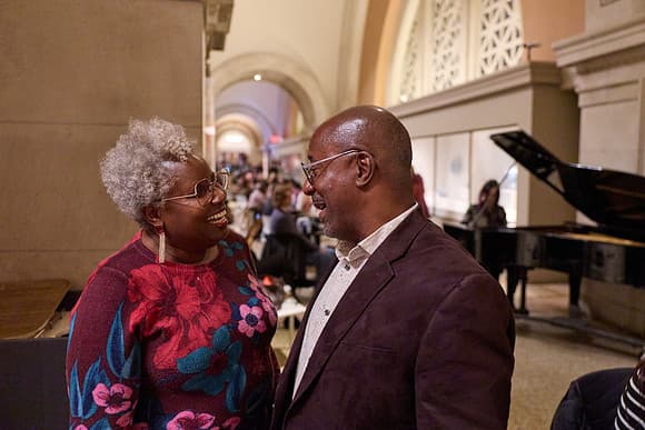 Two people face each other and smile. The individual on the left is brown skinned, feminine presenting with gray curly hair, glasses, dangly silver earrings. This person wears a burgundy dress with large floral motifs in red, pink, and blue. The individual on the right is brown skinned, masculine presenting, bald and wearing glasses. They are wearing a cream button down shirt with a maroon blazer. In the background, a feminine presenting person is playing the piano to a seated group.