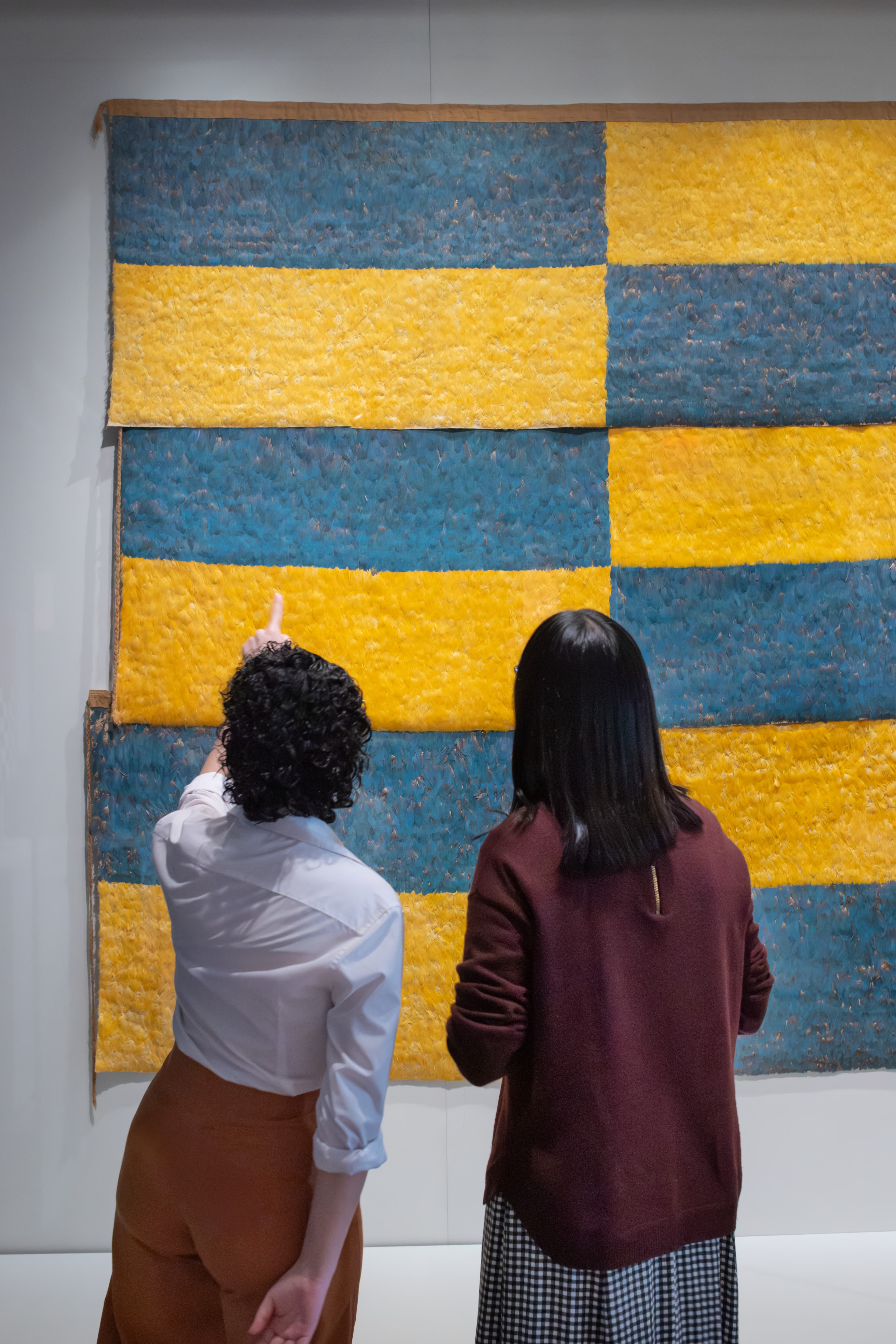 Two visitors look at panels of feathers on the wall. The panels are blue and yellow and form a checkered pattern.