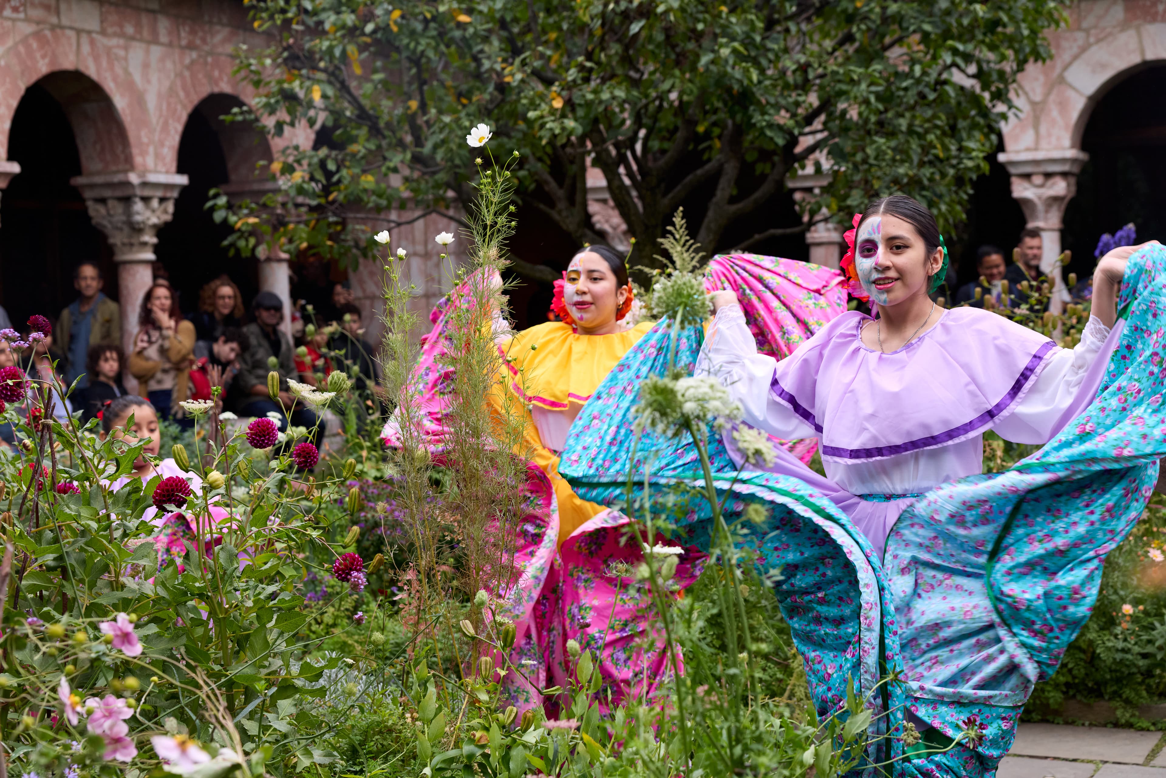 Two young women dance in a garden. They are wearing colorful dresses and half of their faces are painted.
