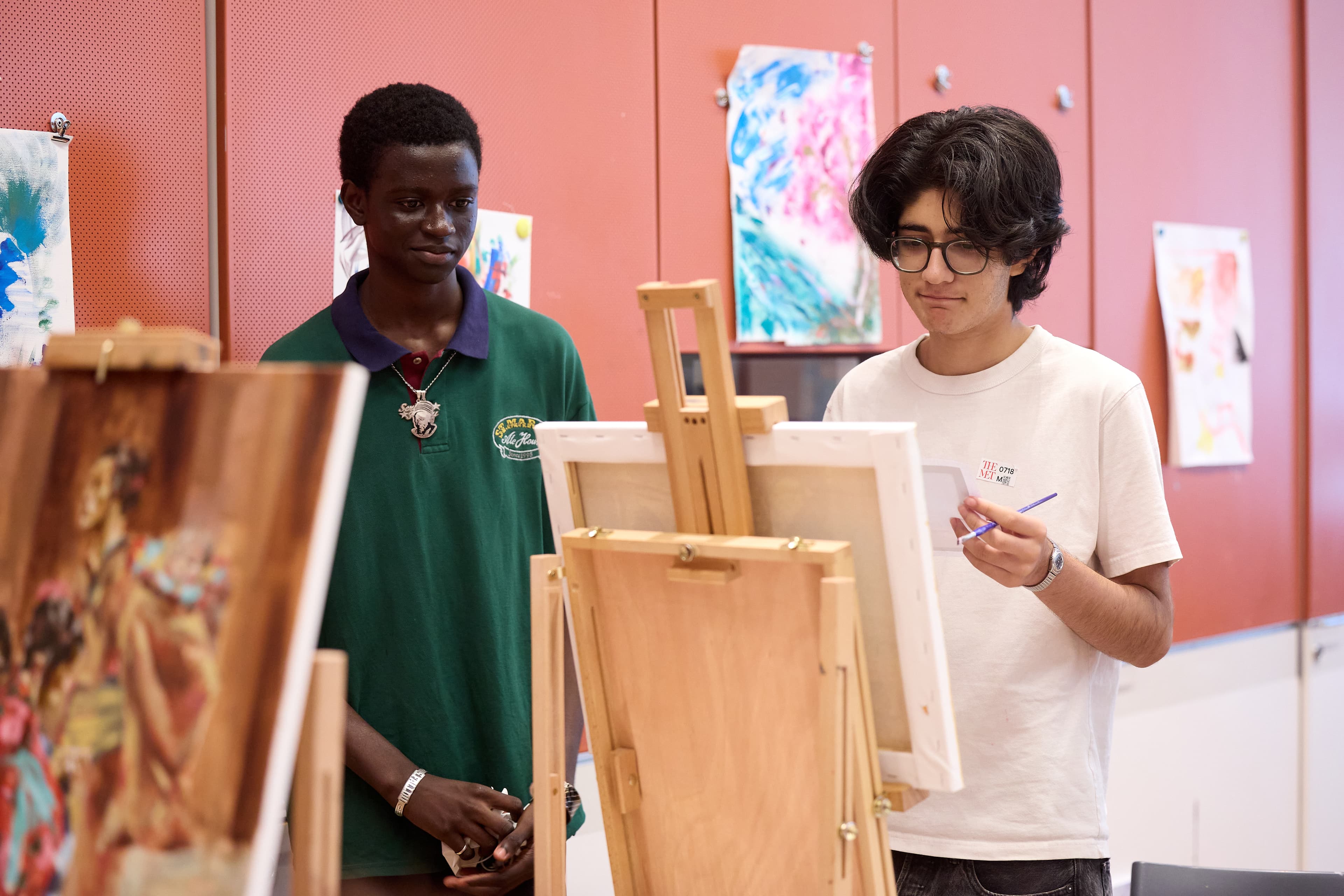 Two teens are looking intently at an easel with a canvas on it. There are other paintings hung up on the wall behind them.