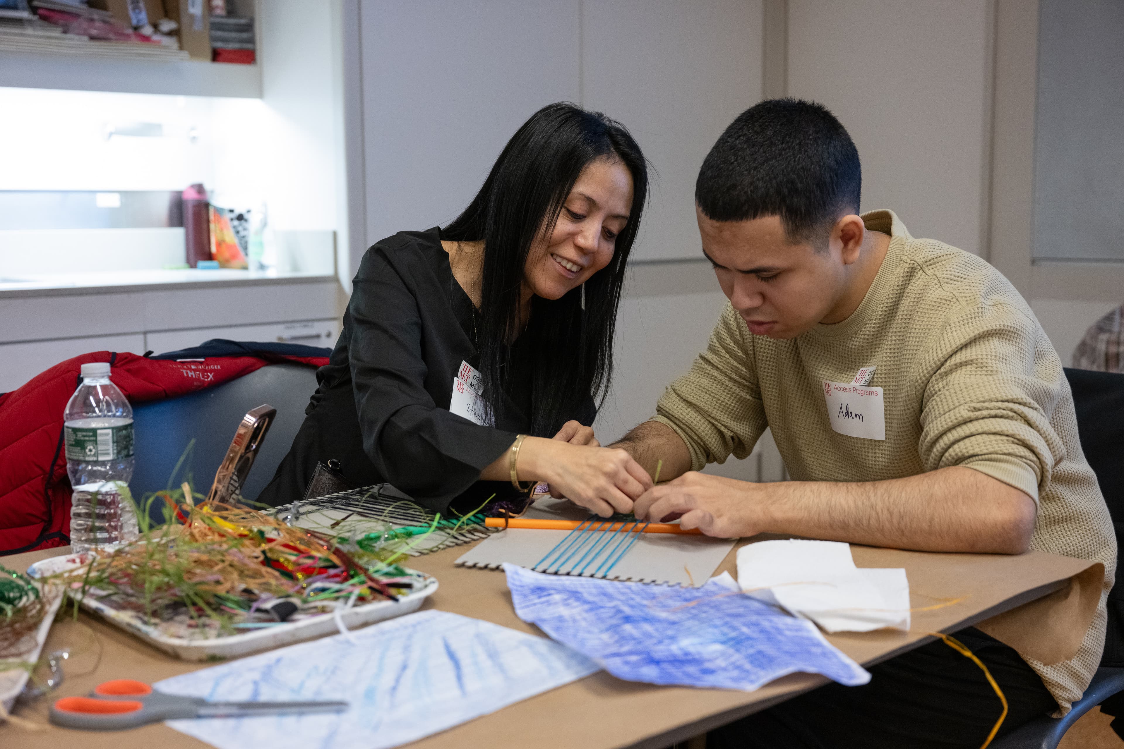 Two visitors sit at a table in a studio classroom at The Met as they work on a weaving project with yarn and a cardboard loom. Other art materials surround them at their table.