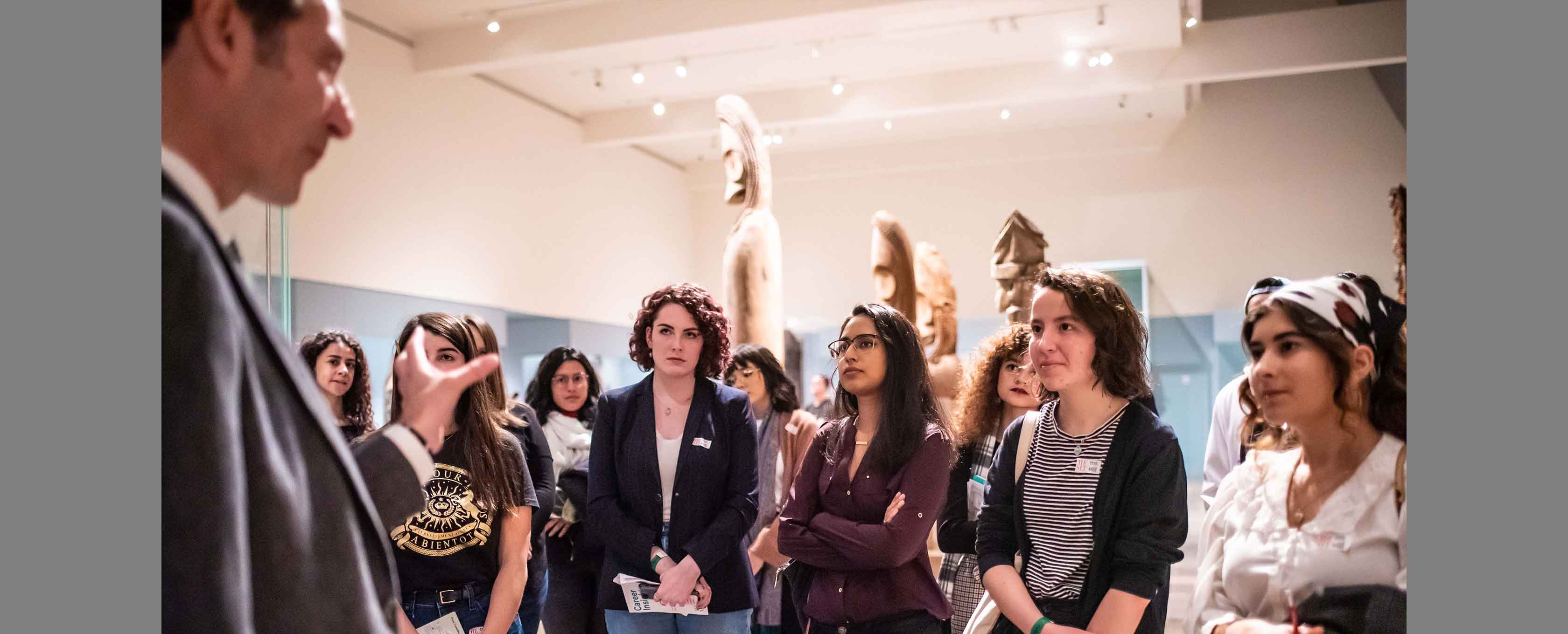 A group of nine college students in casual clothes stand in a gallery listening to someone speaking mostly off-camera, in the foreground.